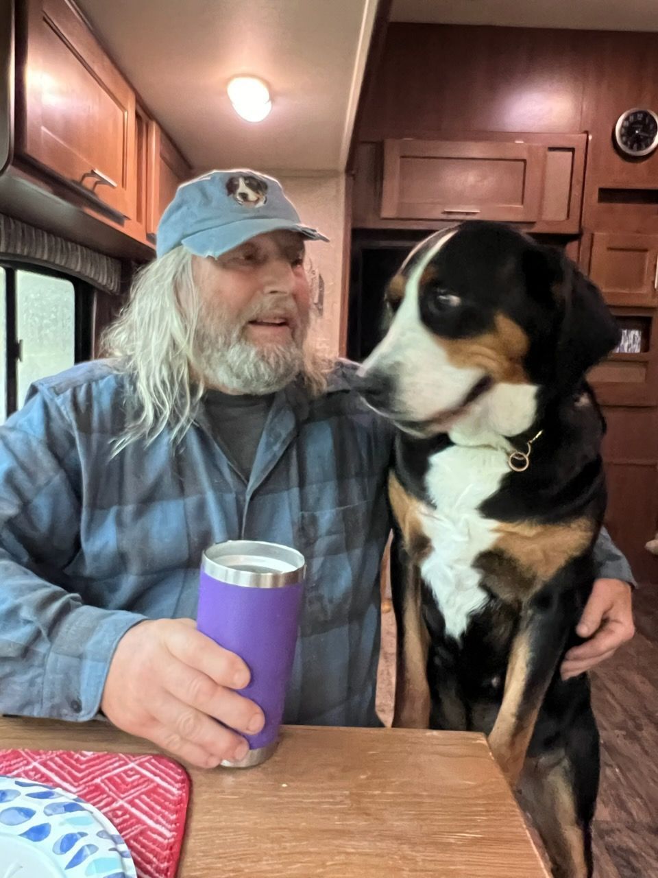 Person in a blue cap sits at a table with a black, white, and tan dog in an RV kitchen.