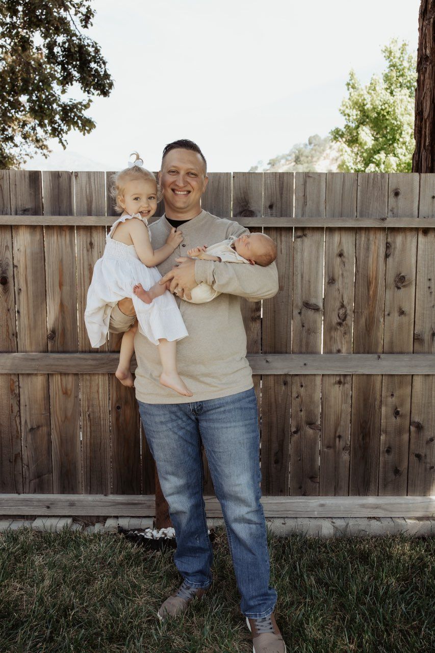 Person standing outdoors holding two small children in front of a wooden fence