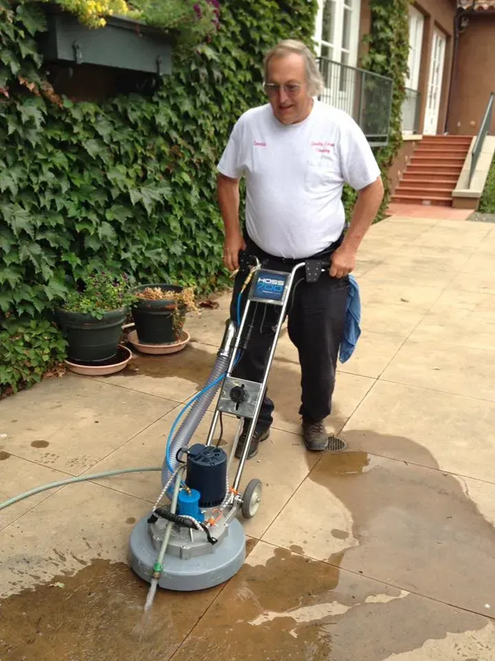 Person using a floor-polishing machine on a wet outdoor walkway beside a vine-covered wall