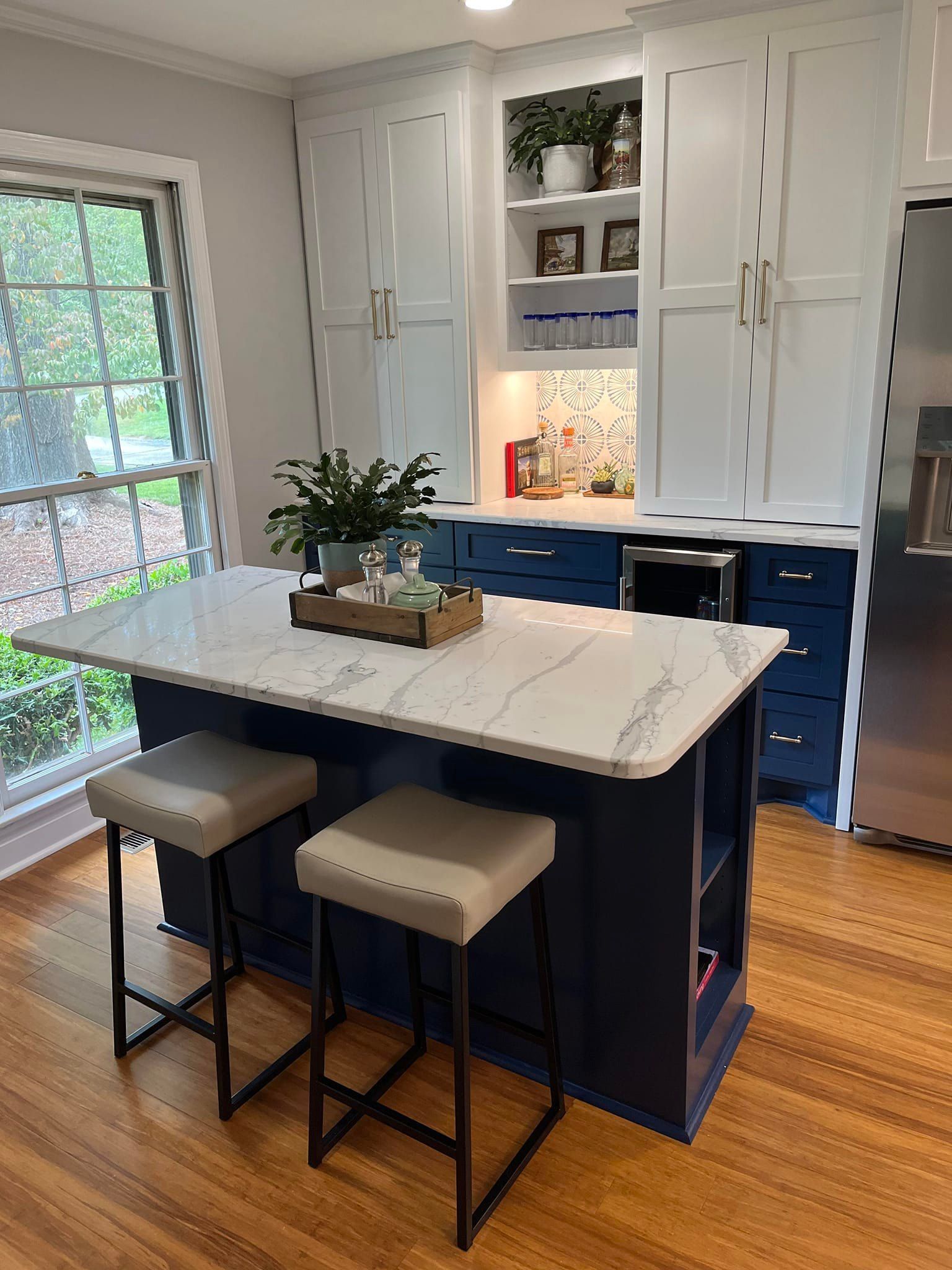 A kitchen island with a white marble top and navy base features two taupe barstools in a room with white cabinets.