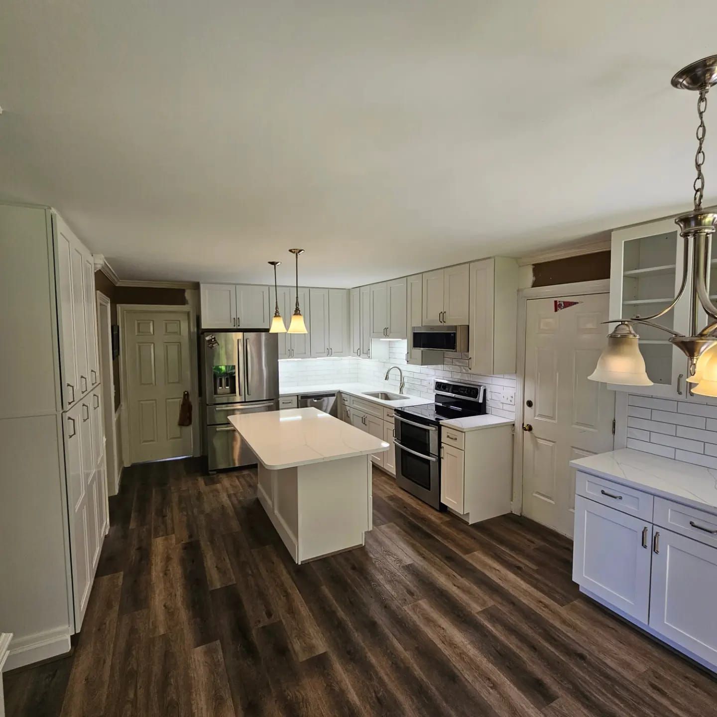A bright, modern white kitchen with wood-look floors, a central island, stainless steel appliances, and pendant lighting.