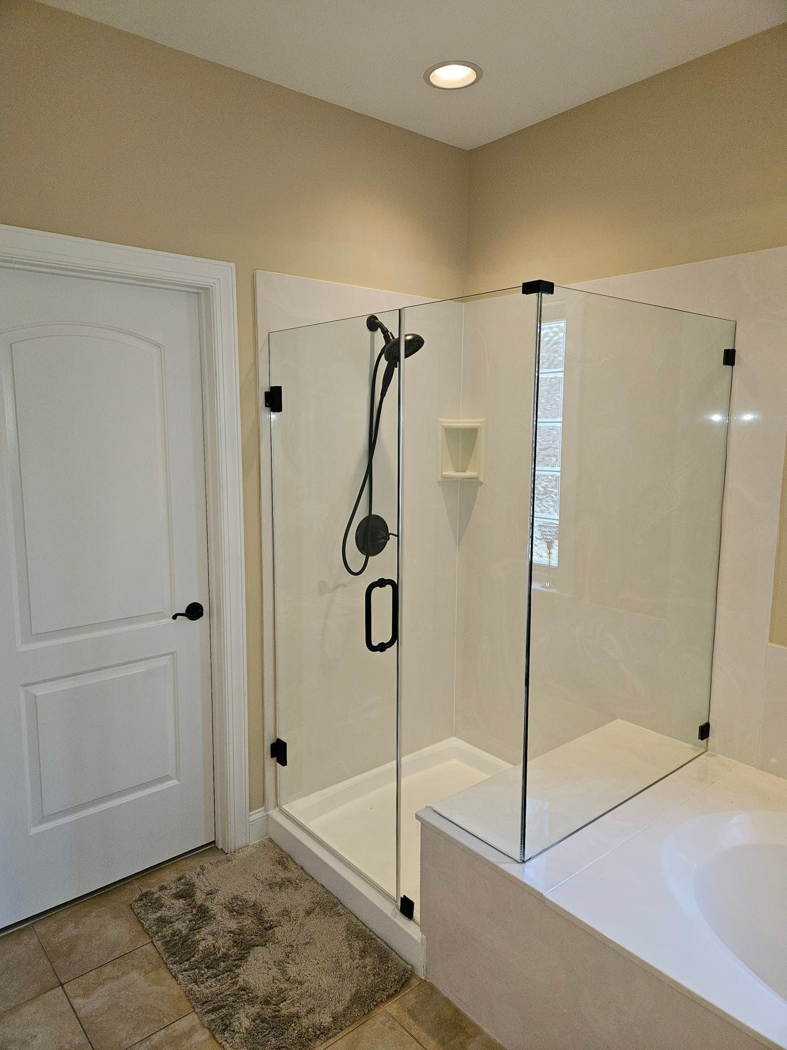A modern bathroom features a glass walk-in shower with black fixtures next to a white door and light-colored tile floors.
