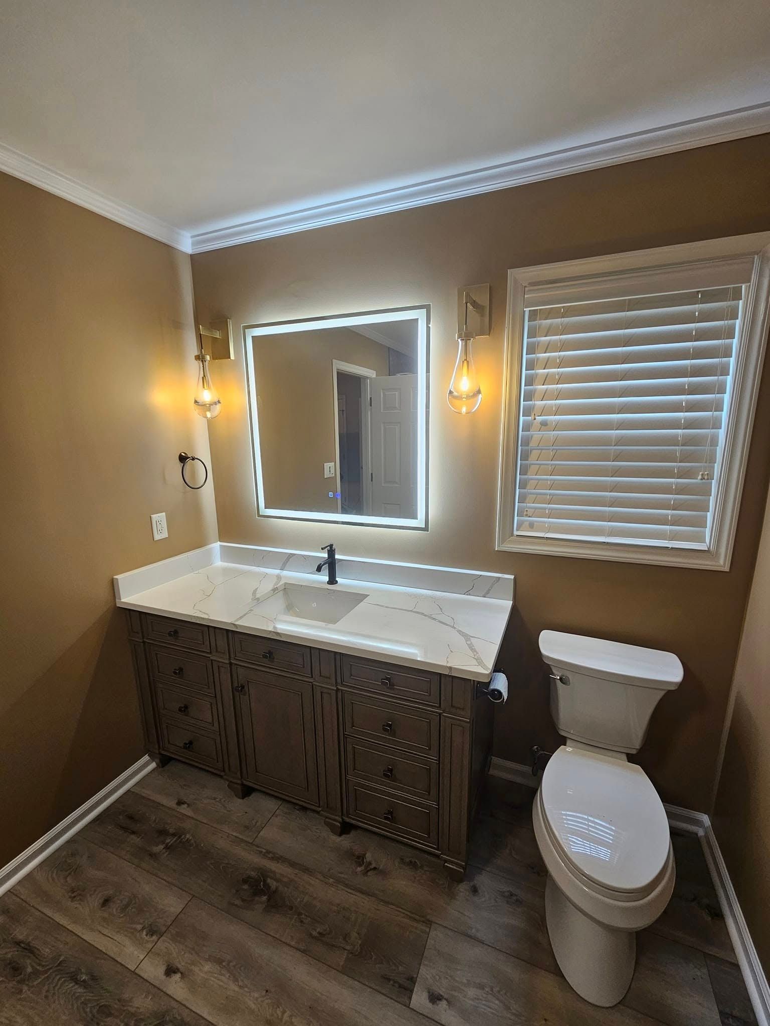 A bathroom with a brown vanity, white countertop, lighted square mirror, two wall sconces, a toilet, and wood-look floors.