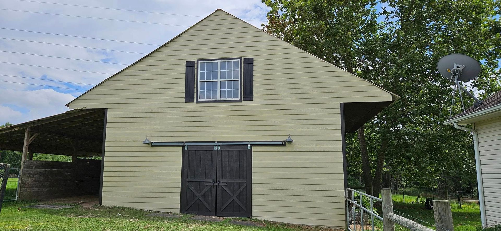 A two-story, light yellow barn with a dark brown sliding door, upper window, and an attached open-sided shelter.