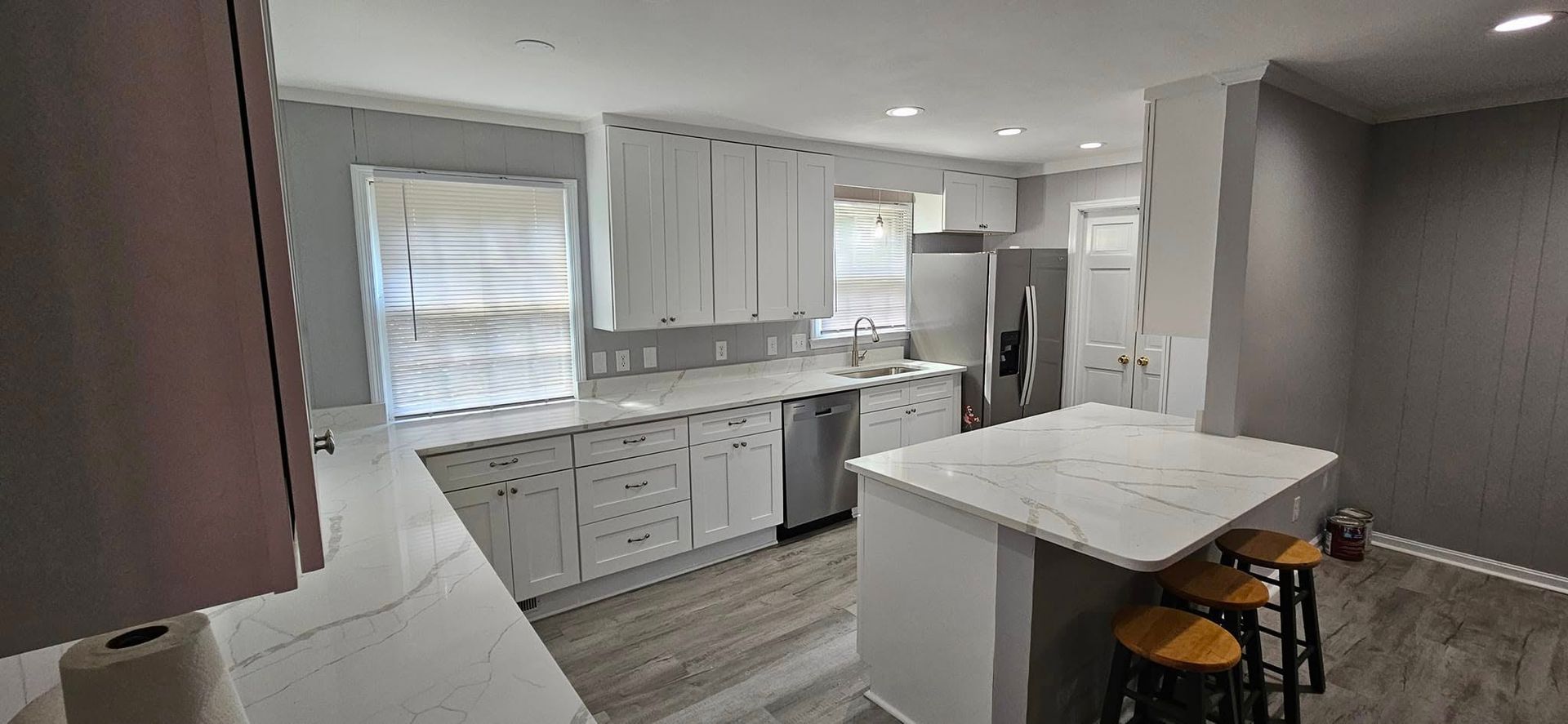 A modern kitchen with white cabinets, light countertops, a center island with three stools, and stainless steel appliances.