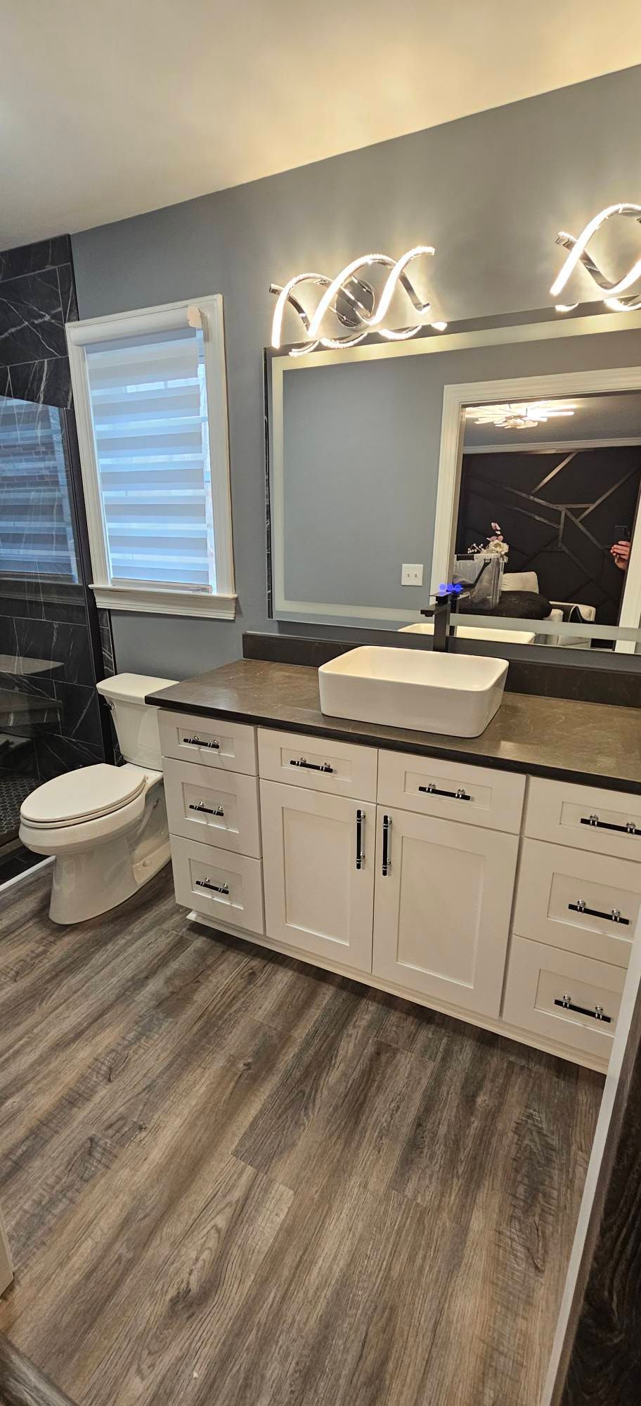 A bathroom with white cabinets, a square vessel sink, a large mirror with modern lights, and wood-look flooring.