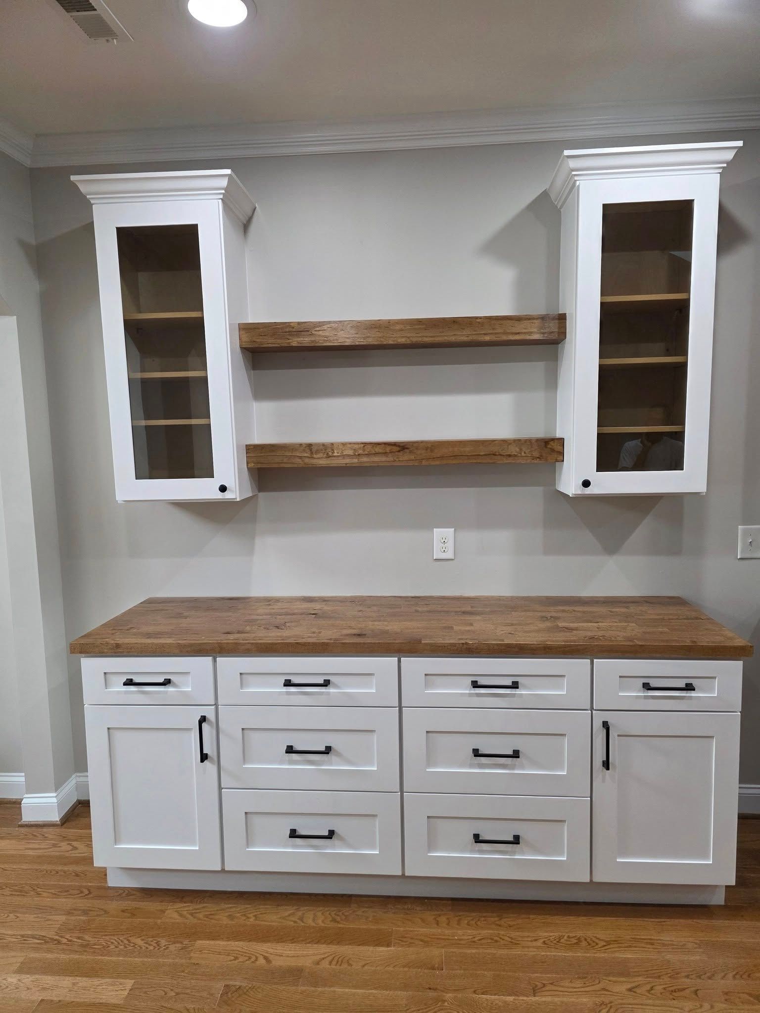 White kitchen cabinetry featuring a butcher block countertop, two glass-front upper cabinets, and two floating wood shelves.