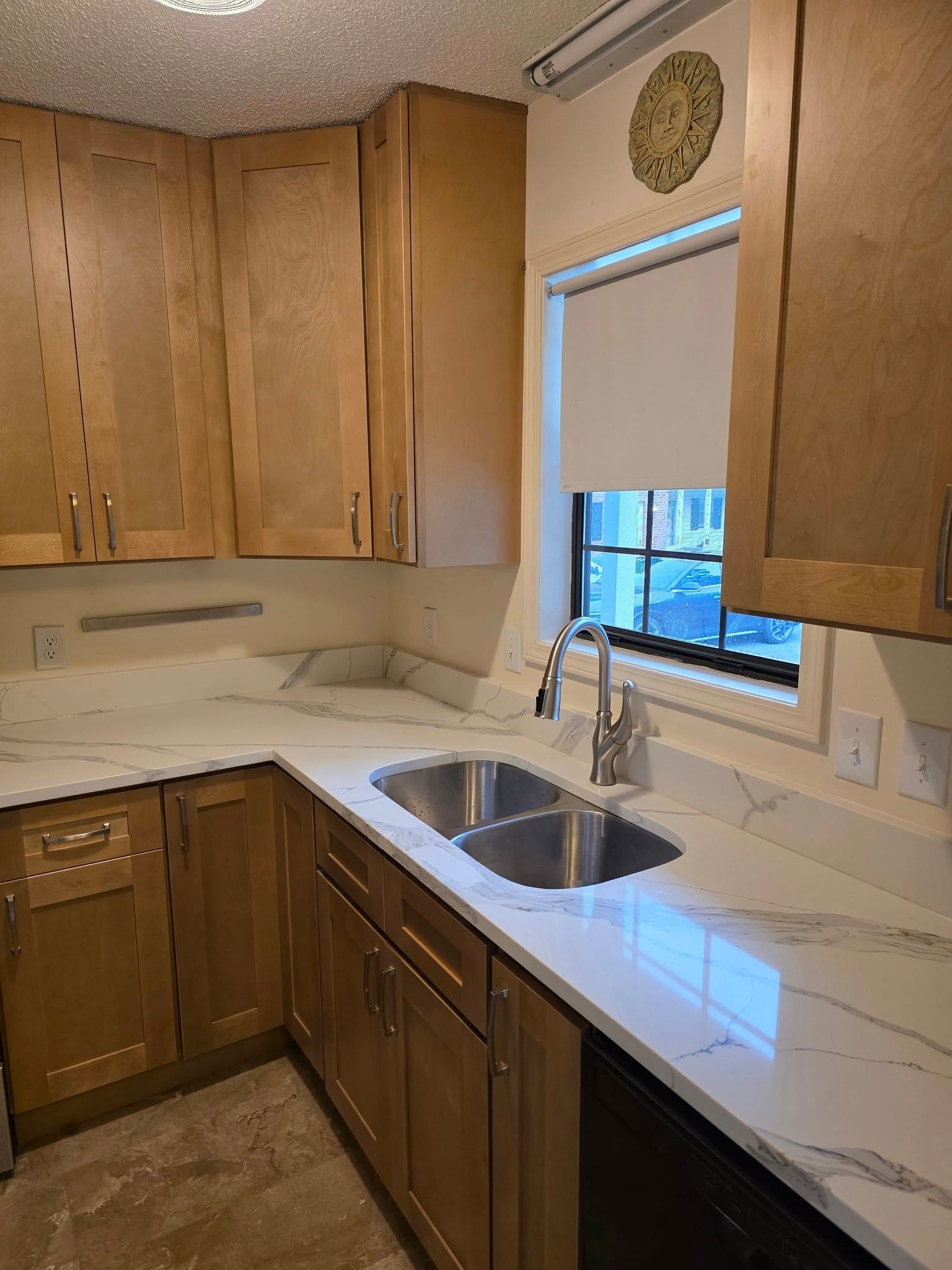 A kitchen corner featuring wood cabinets, white marble-patterned countertops, a double basin sink, and a window with a shade.