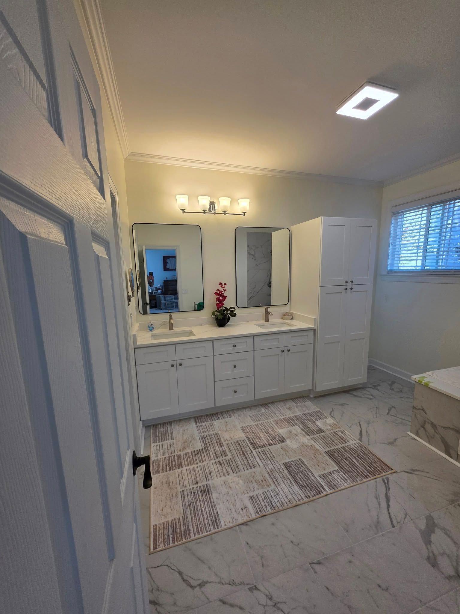 A bright, modern bathroom with a white double vanity, rectangular mirrors, a tall cabinet, and light marble-patterned tile.