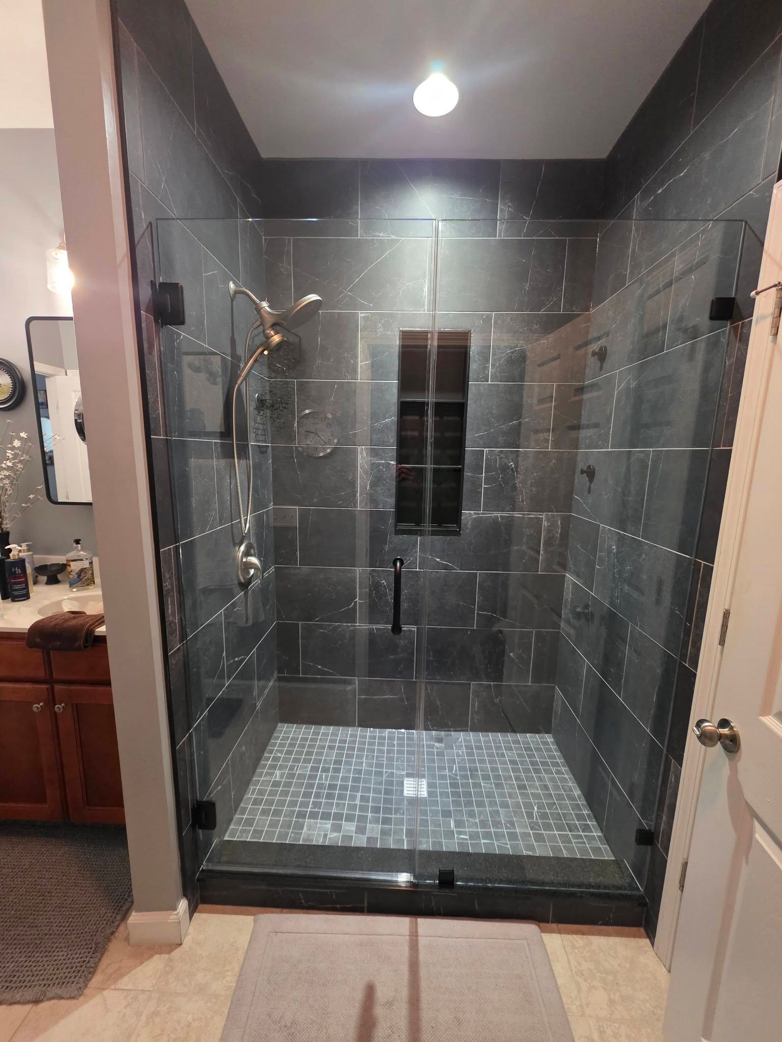 A walk-in shower with dark gray stone tile walls, a mosaic tiled floor, a glass door, and a built-in shelf.