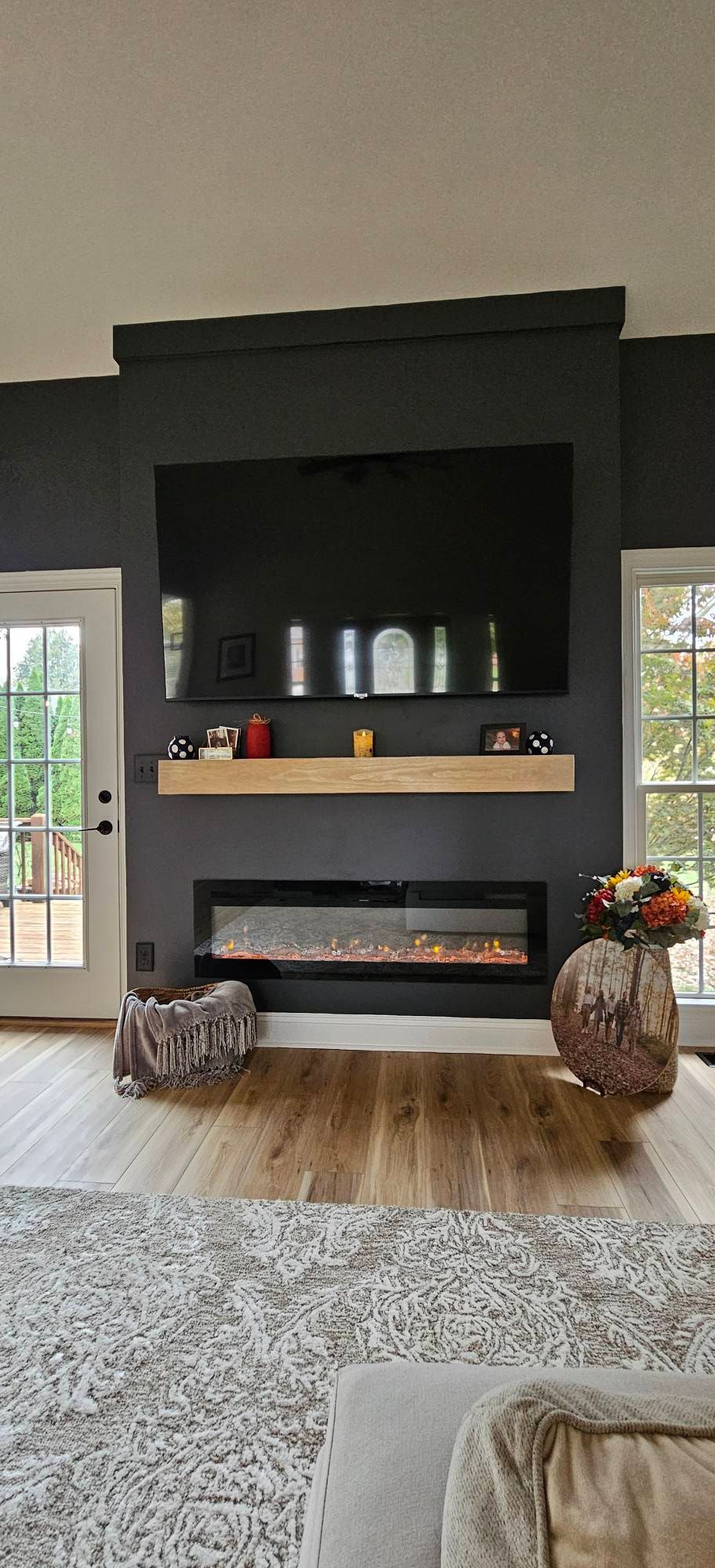 A dark accent wall with a mounted flat-screen TV above a light wood mantel and a long, linear electric fireplace.