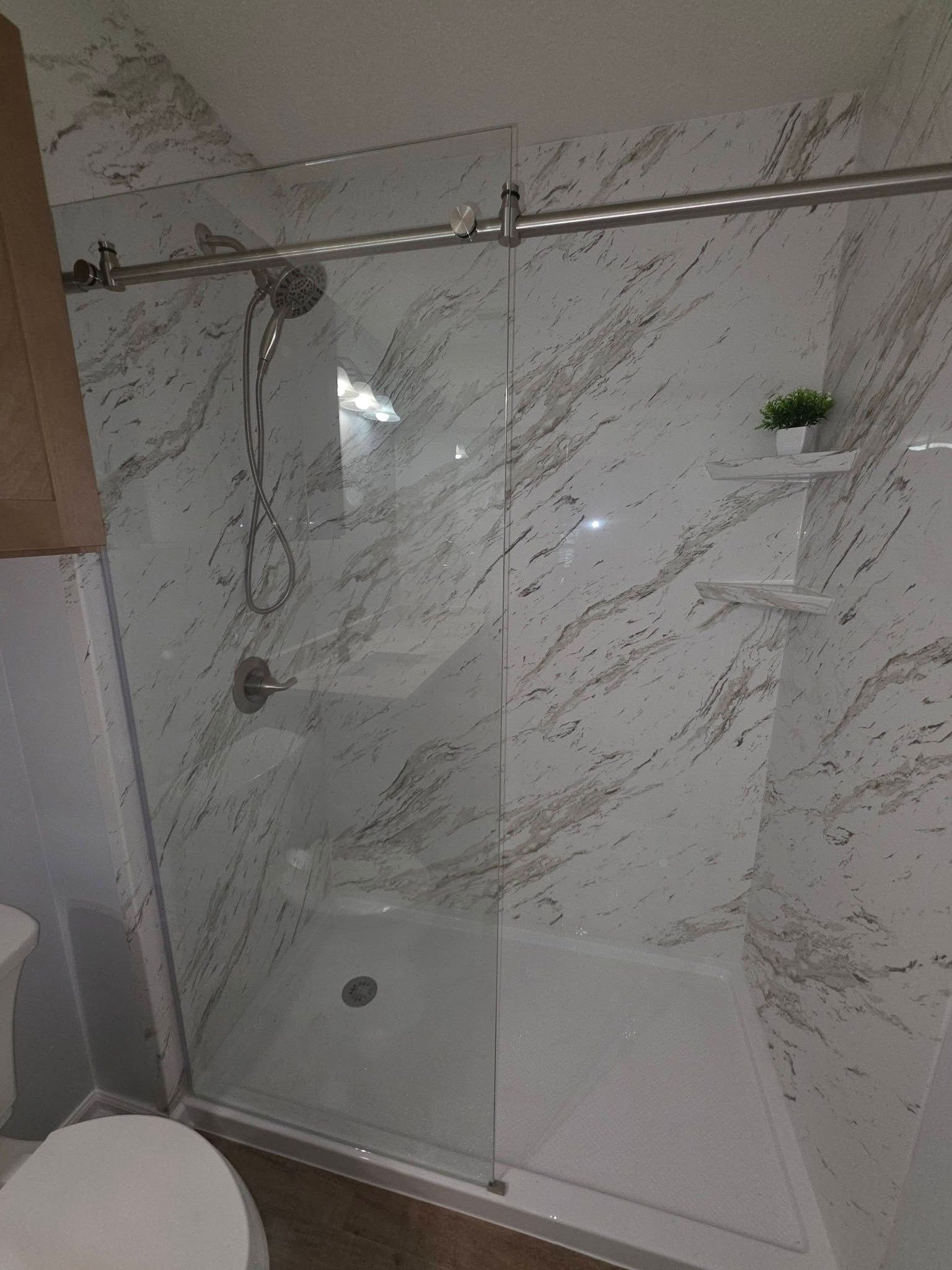 A walk-in shower with white and grey marble-patterned walls, a sliding glass door, and two built-in corner shelves.
