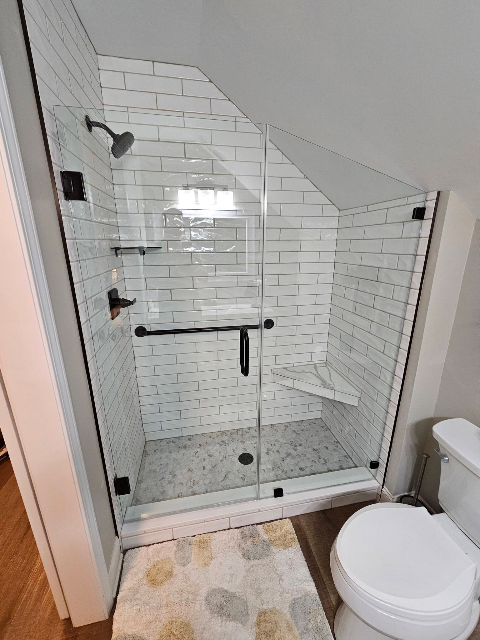 A walk-in shower with white subway tile, a glass door, a corner bench, and a grey patterned floor next to a toilet.