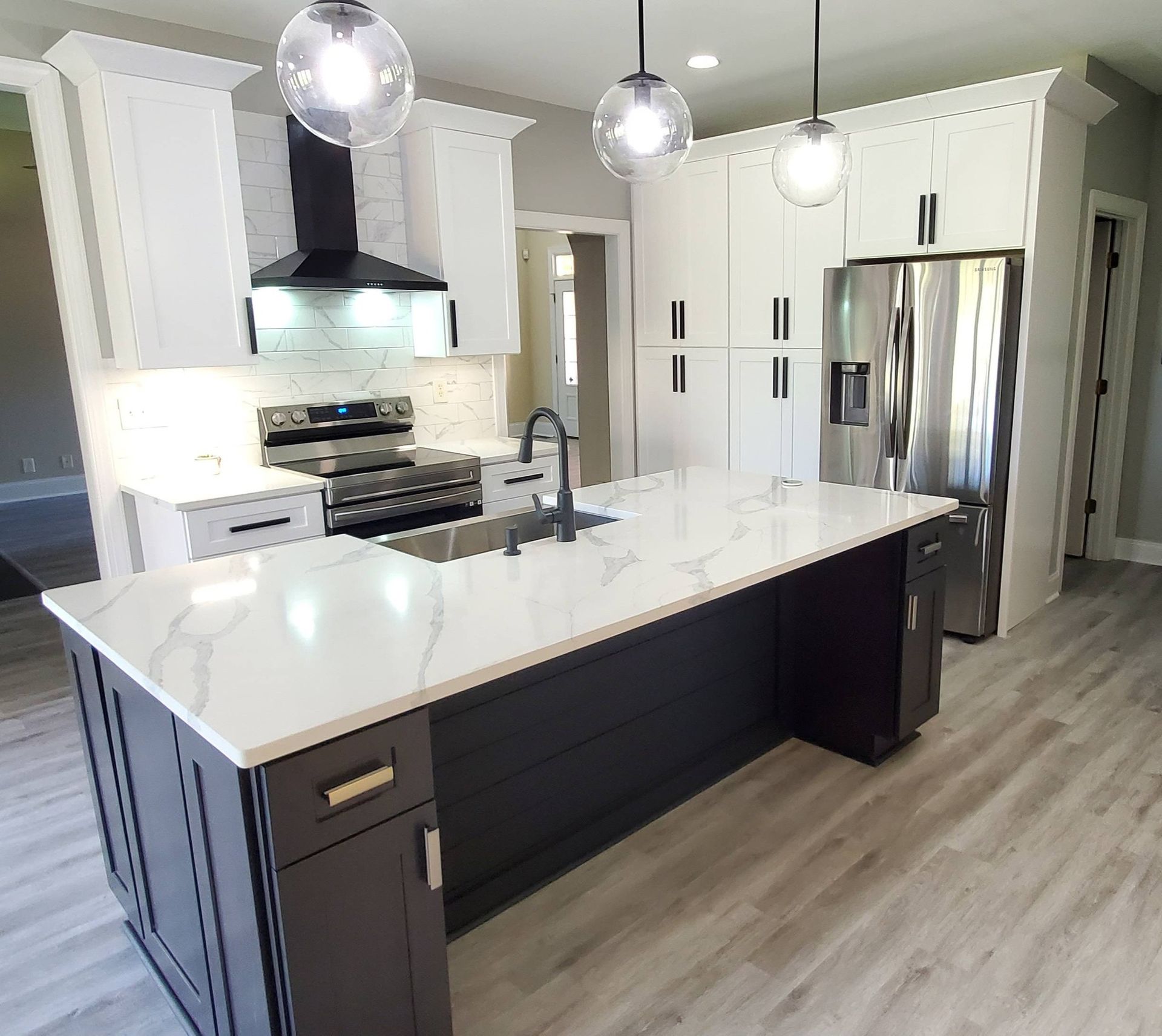 Modern kitchen with a dark-colored island, white cabinets, stainless steel appliances, and wood-look flooring.