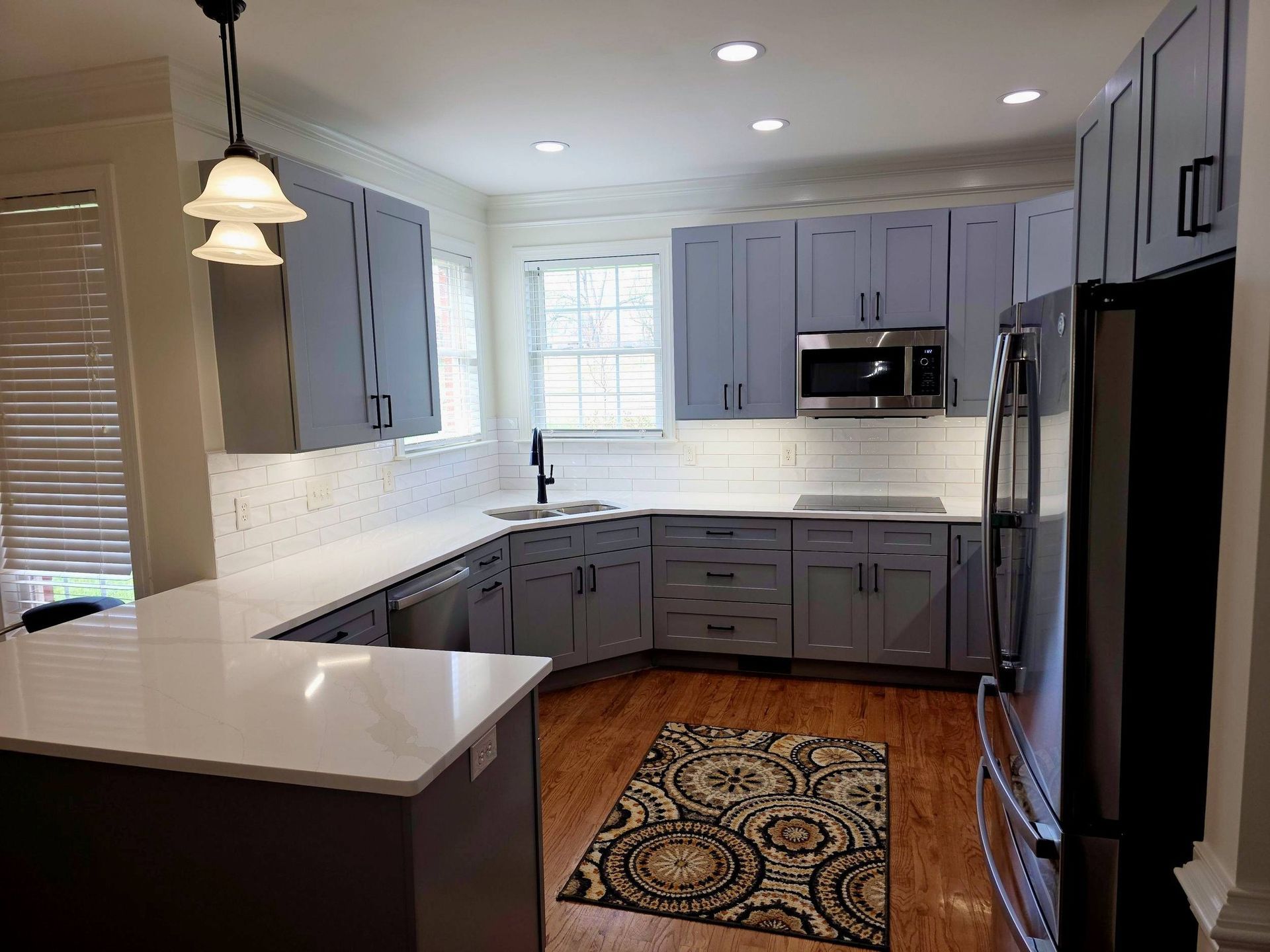 A modern kitchen with grey cabinets, white countertops, stainless steel appliances, and a patterned rug on hardwood floors.