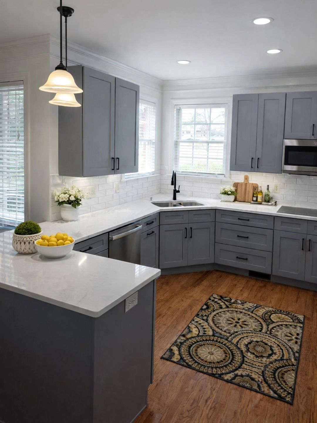 Modern kitchen with gray cabinets, white countertops, marble backsplash, a patterned rug, and two hanging pendant lights.