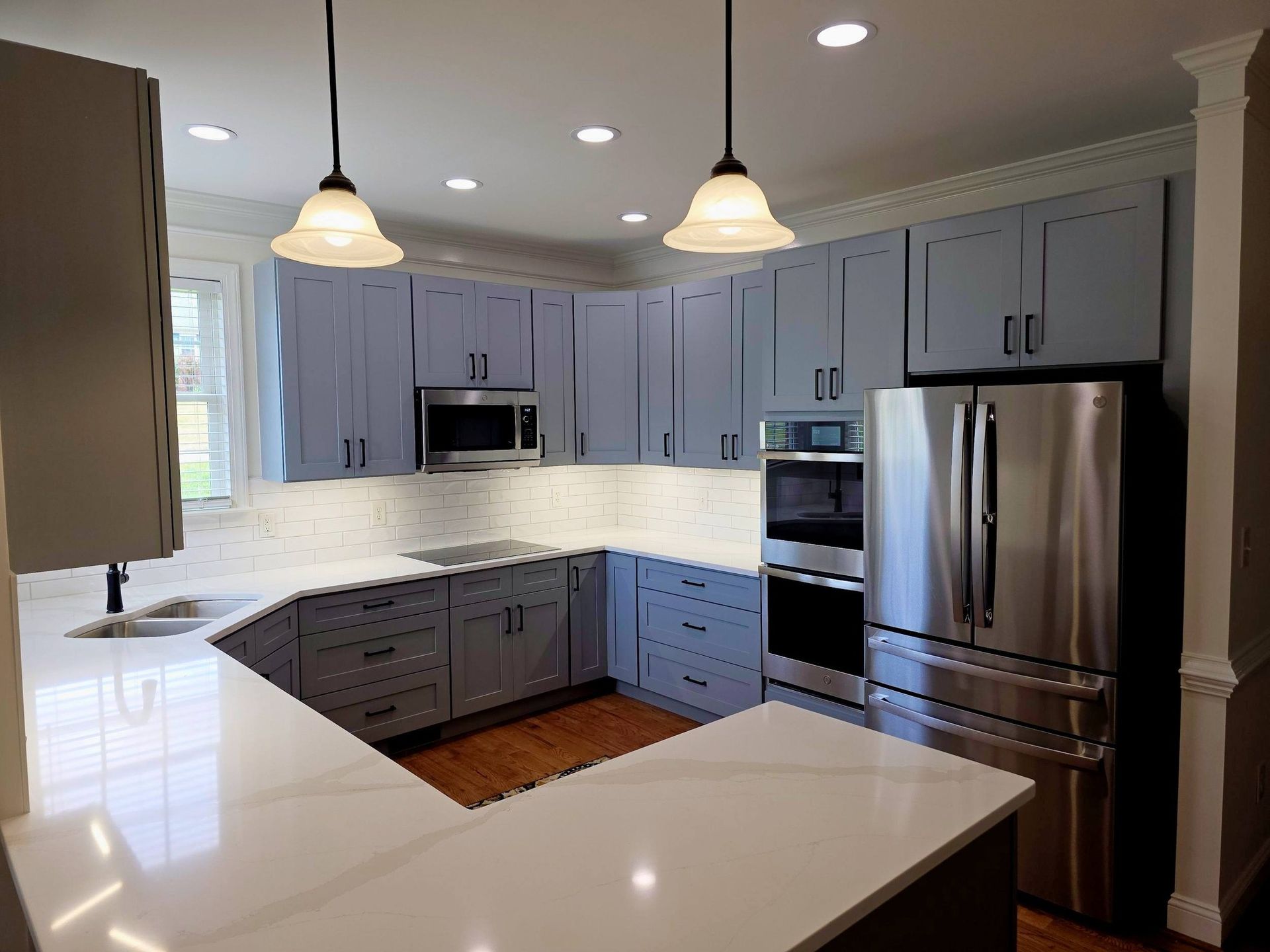 A modern kitchen featuring U-shaped white countertops, light blue cabinets, stainless steel appliances, and two pendants.