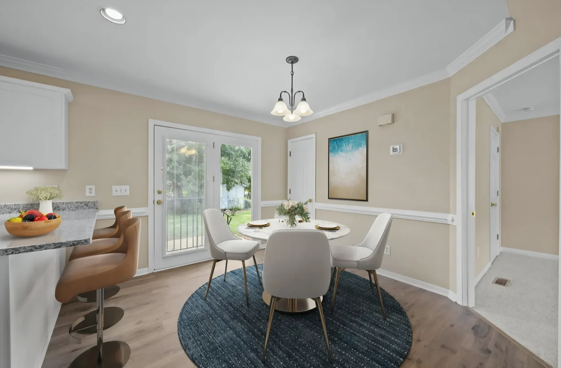 A dining area with a round table, four white chairs on a blue rug, and kitchen bar stools, near a glass patio door.