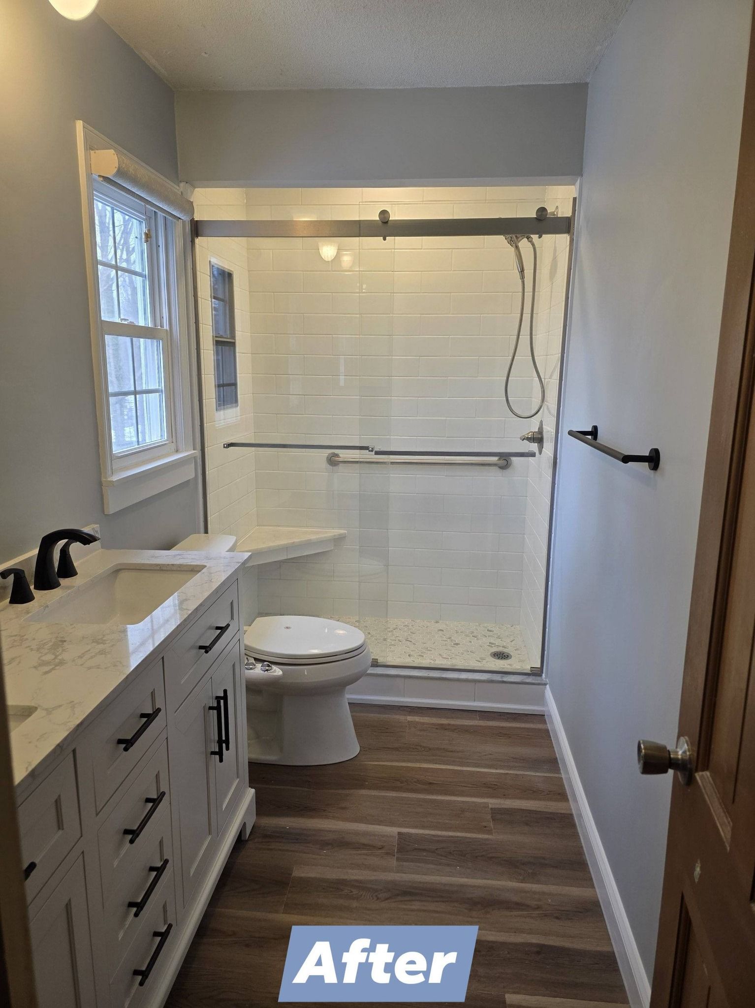 A renovated bathroom with a white vanity, wood-look flooring, and a glass-enclosed walk-in shower with tile walls.