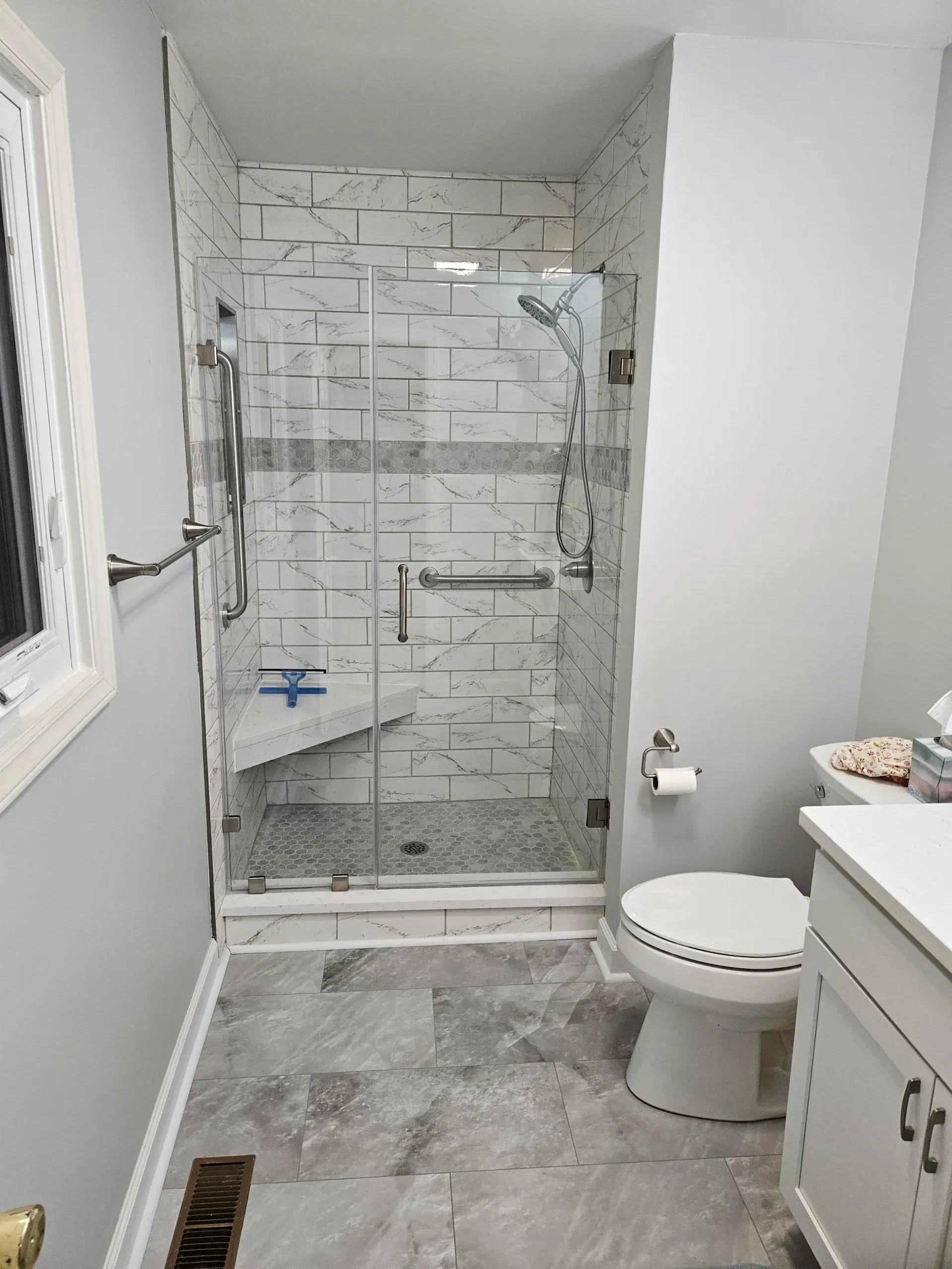A bright, modern bathroom featuring a walk-in glass shower with marble-look tiles, a built-in bench, and a white toilet.