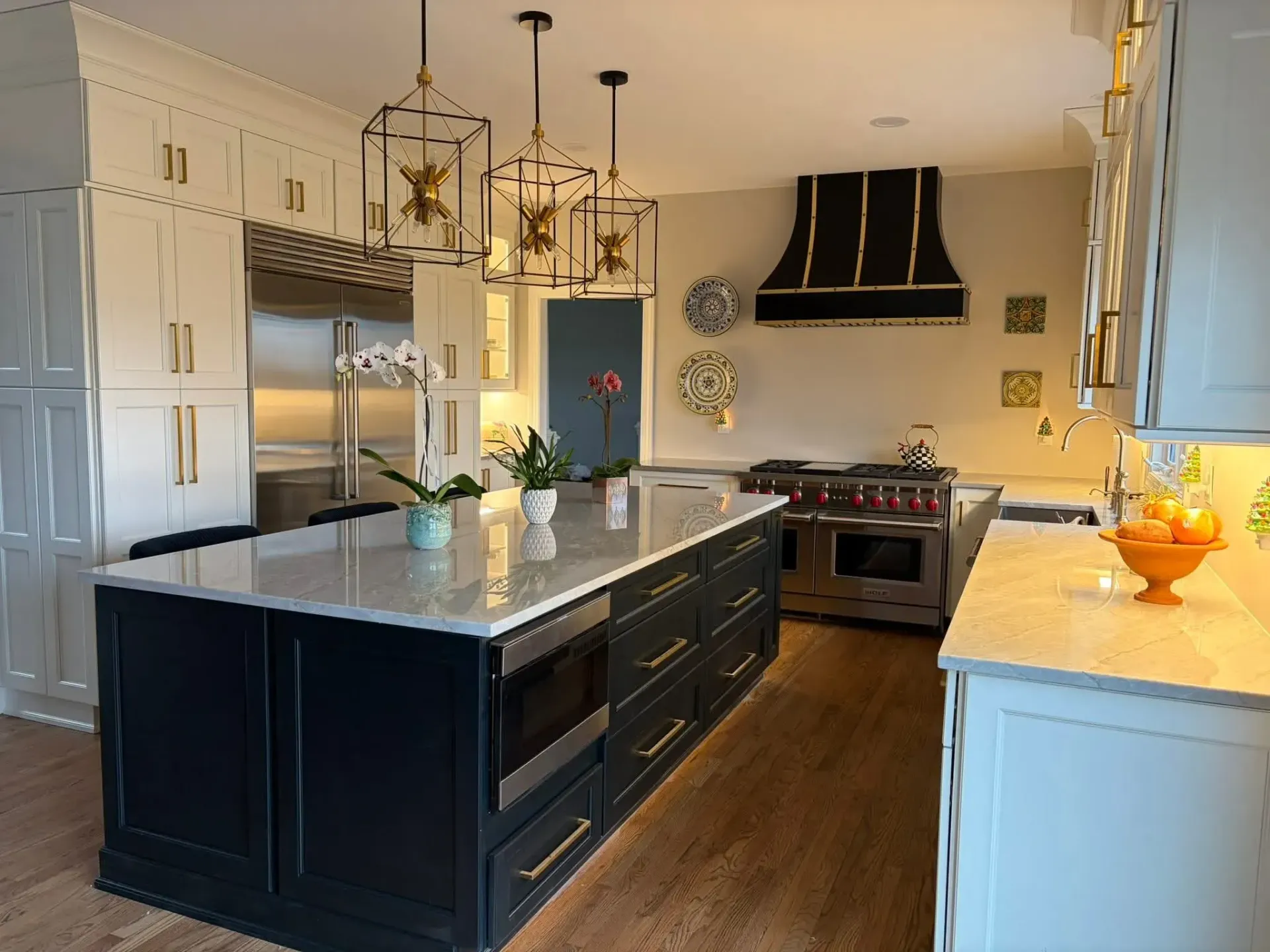 A high-end kitchen with a dark blue island, white cabinets, gold hardware, and three pendant lights over a stone counter.