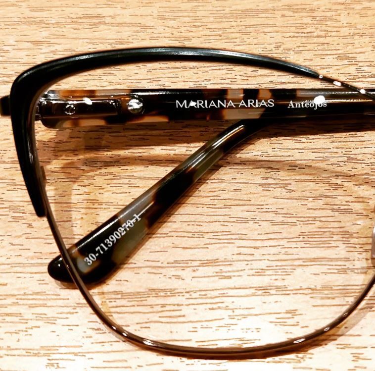 Montura de gafas negra sobre superficie de madera, con lentes transparentes y texto de la marca visible en la patilla.