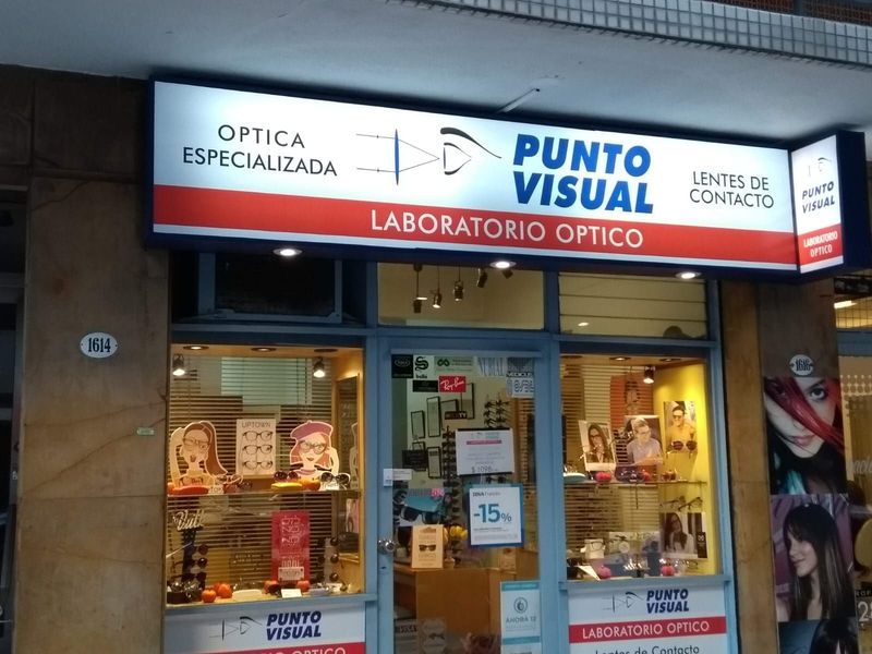 Fachada de la óptica Punto Visual con letrero azul y rojo y expositores de gafas en el escaparate.