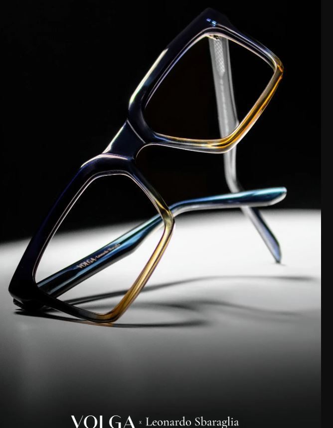 Monturas de gafas geométricas abstractas y brillantes en negro, dorado y azul sobre un fondo oscuro.