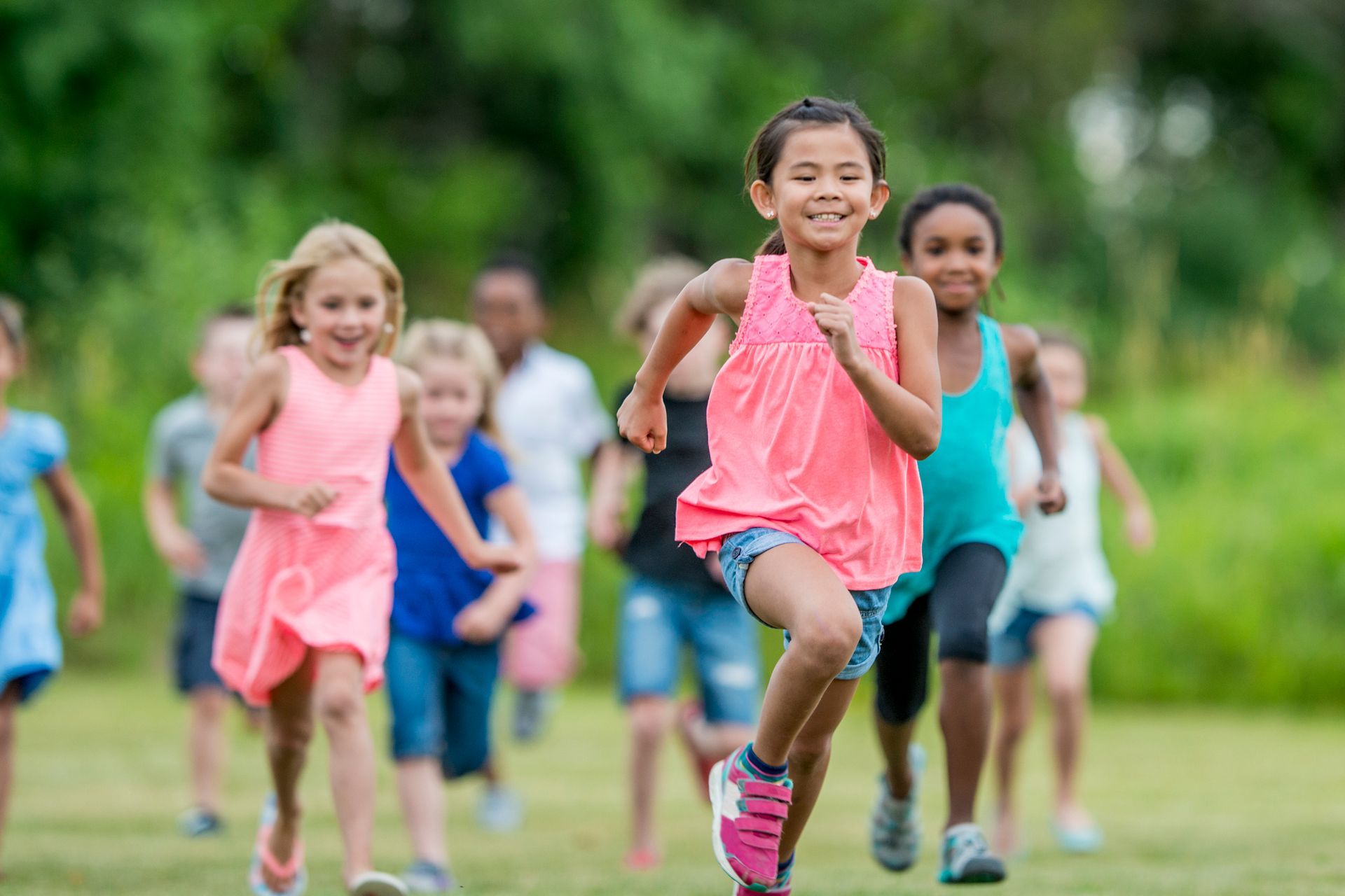 Children running on a grassy field. Girl in pink shirt leads the group, smiling.