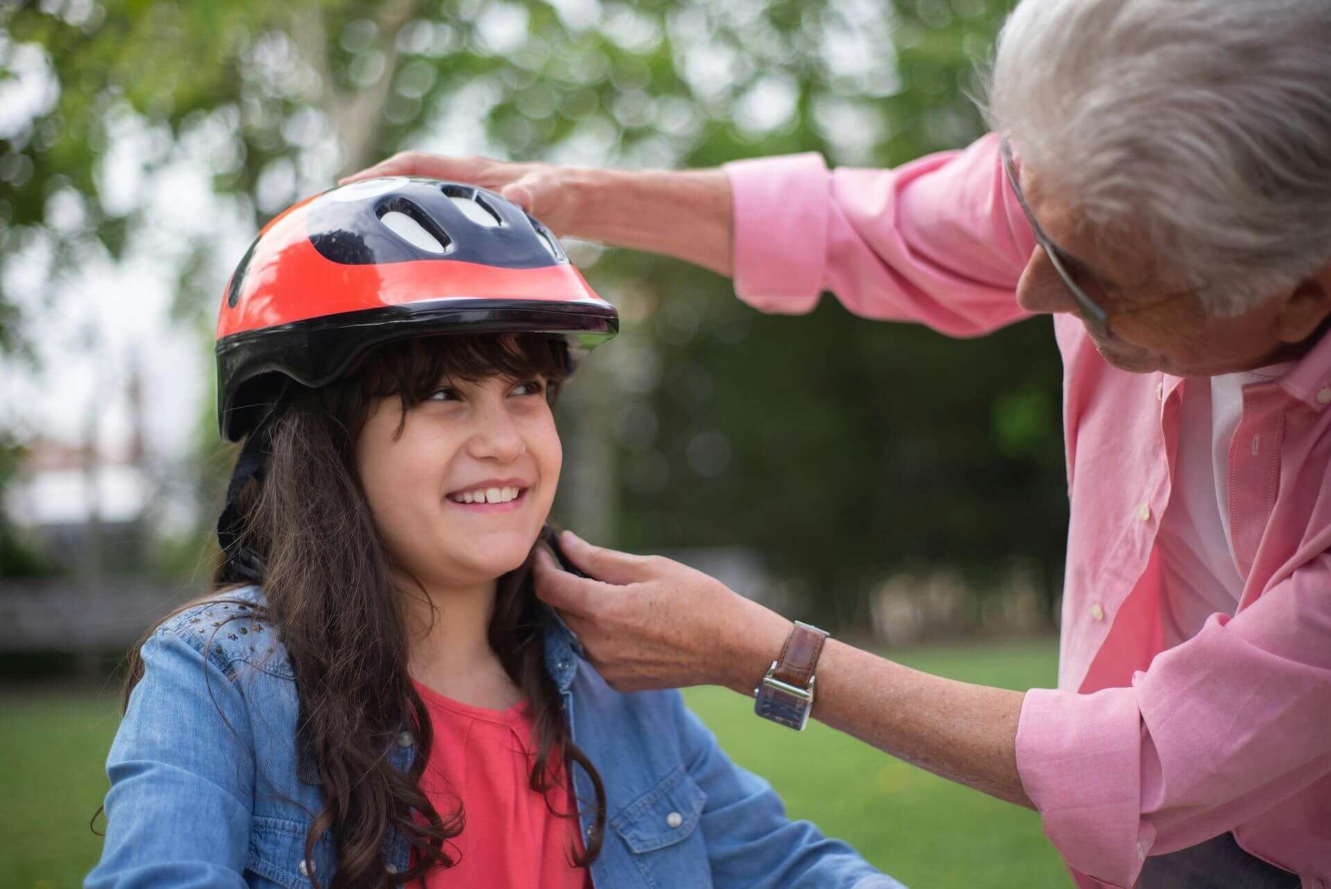 I young girl is outside on her bike and her grandparent is placing a helmet on her. She is smiling.