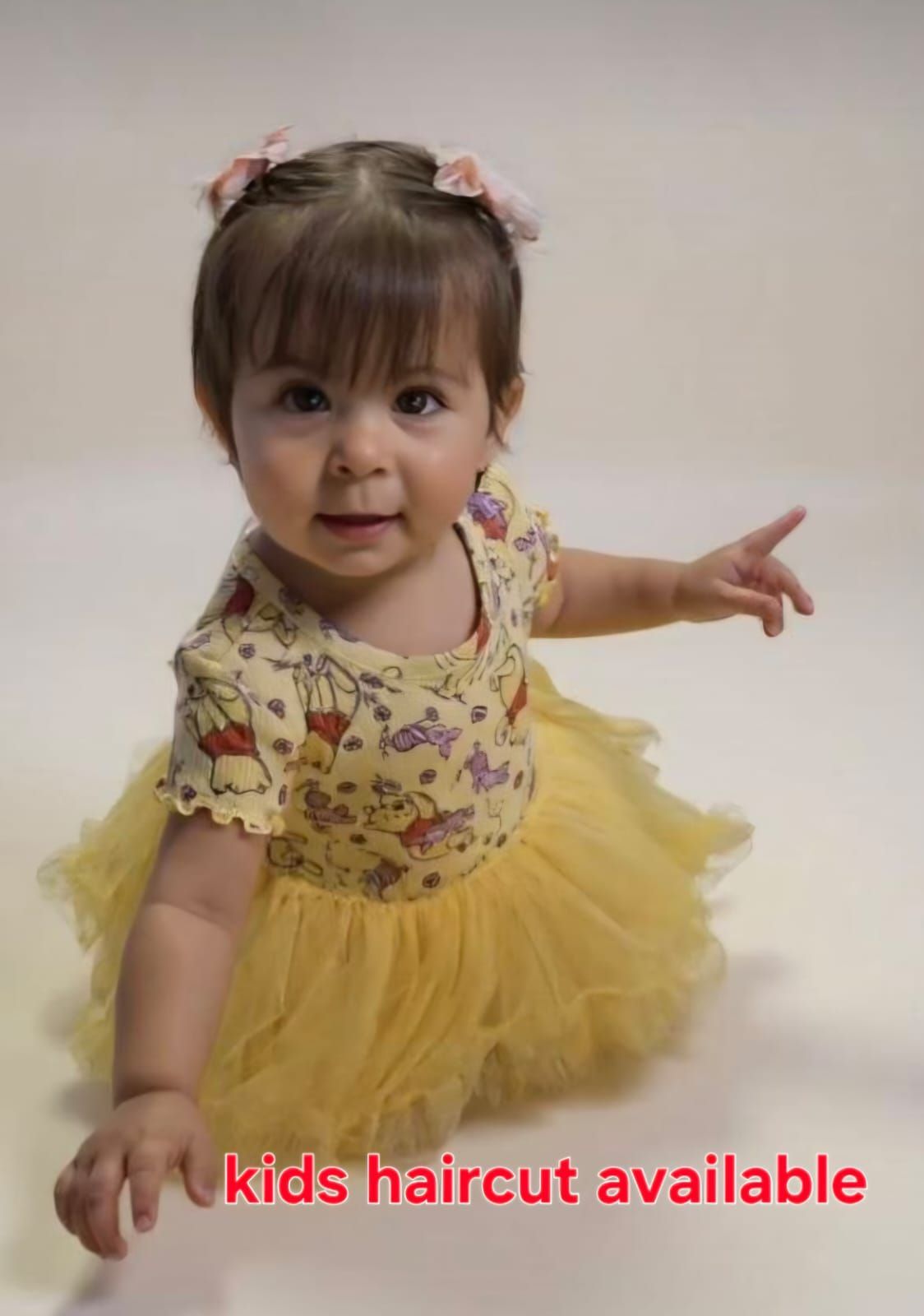Toddler in a yellow dress sitting on a white studio background, reaching out and smiling.