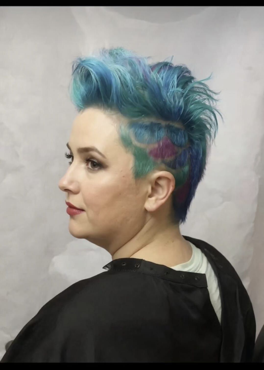 Side profile of a person with blue-green mohawk hair against a plain gray background