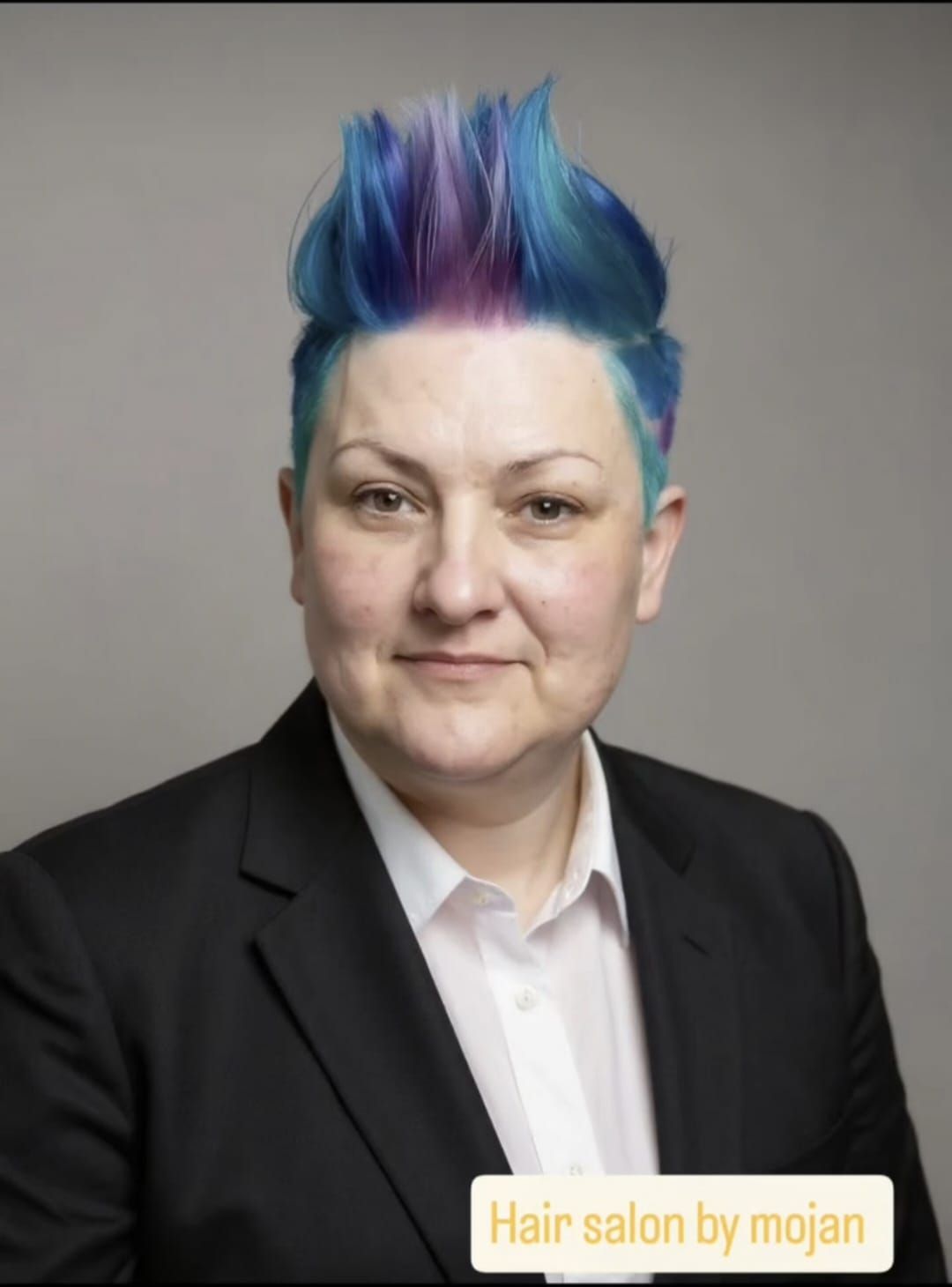 Portrait of a person with blue and purple hair wearing a black blazer and white shirt on a gray background