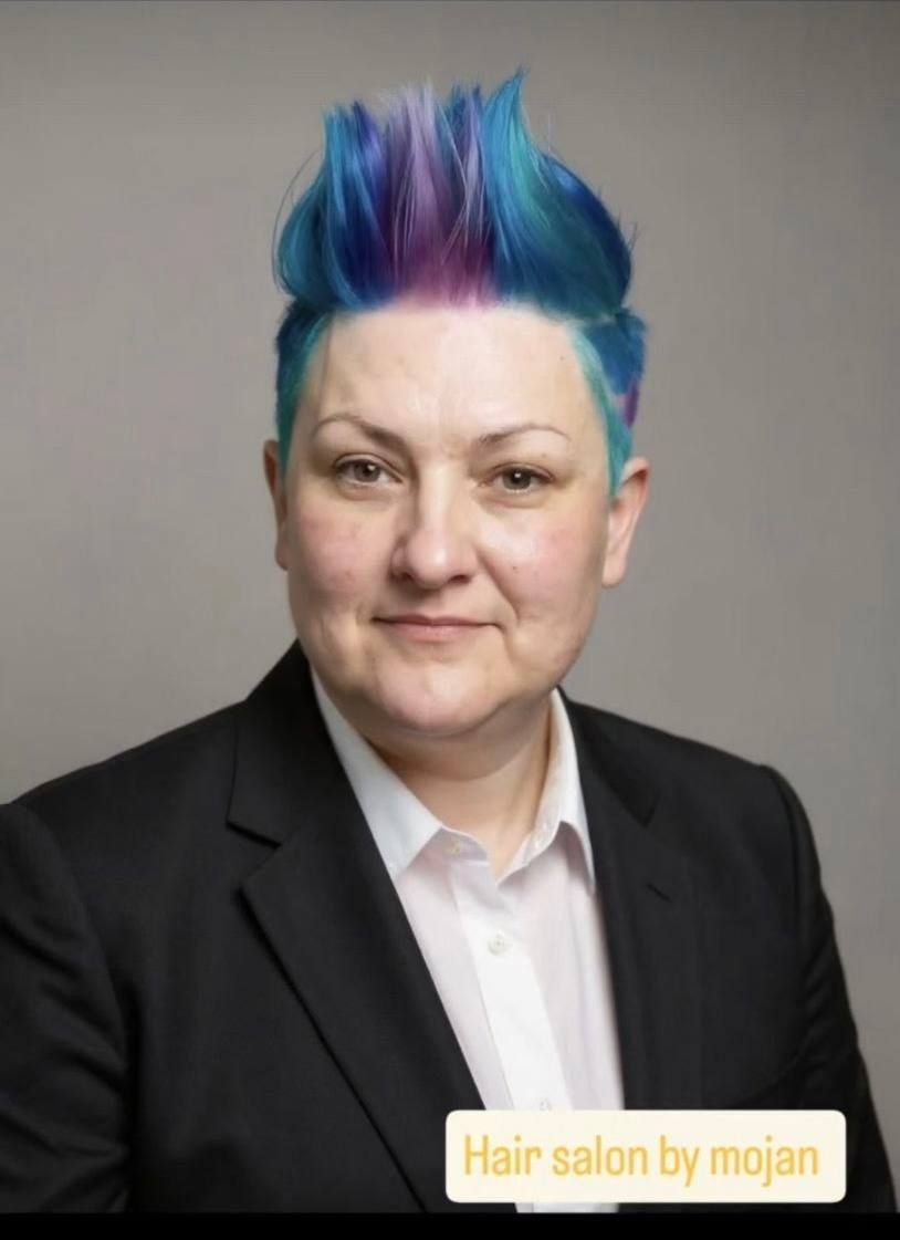 Person with bright blue and purple hair in a black blazer against a gray background.