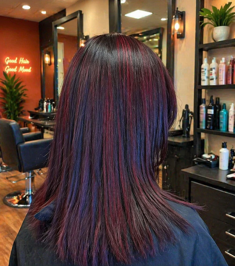 Back view of person with burgundy hair in a salon chair, inside a hair salon with shelves and mirrors.