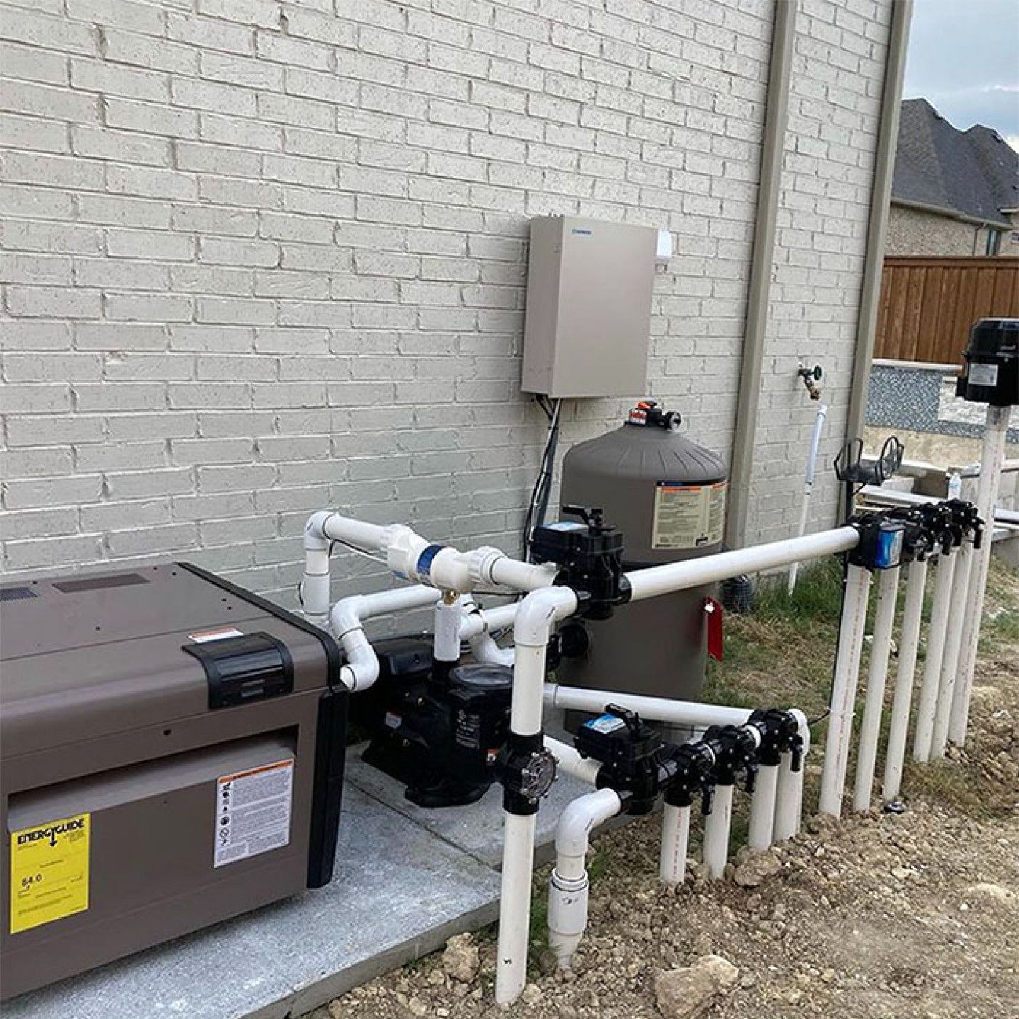 Pool filtration equipment and plumbing mounted beside an outdoor brick wall.