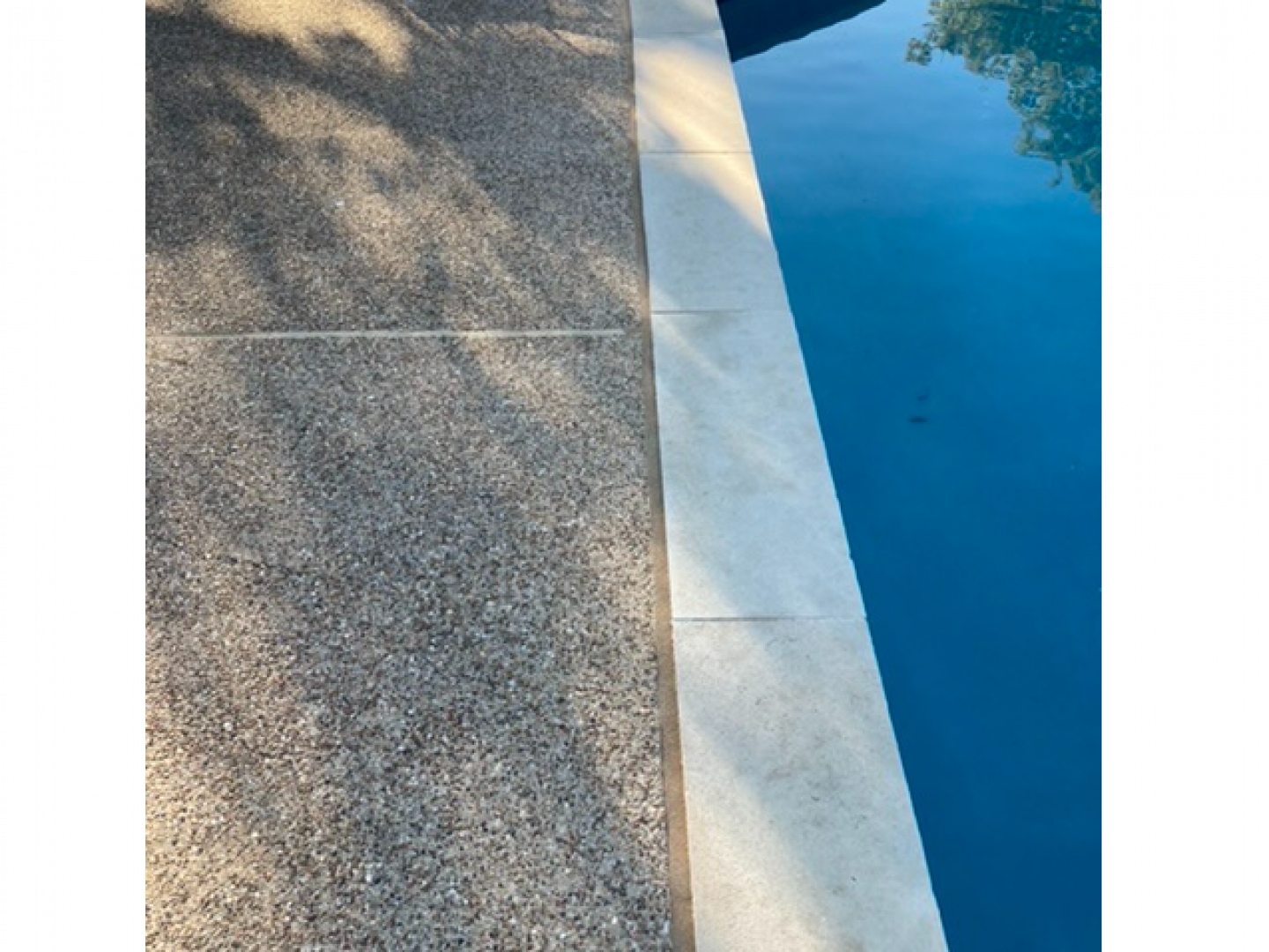 Rough concrete beside a narrow white curb and bright blue pool water with tree shadows