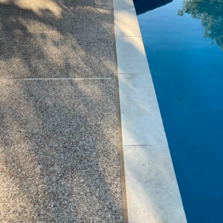 Stone pool deck beside clear blue water with shadows from nearby trees