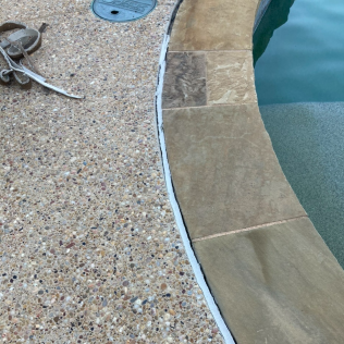 Curved pool edge with tan stone coping beside a pebble-textured deck and turquoise water