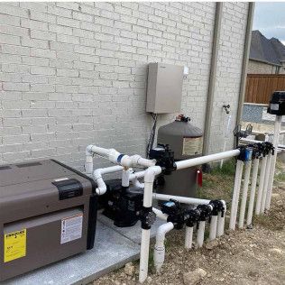 Outdoor pool equipment with gray pumps, white PVC pipes, and a control box beside a brick wall.