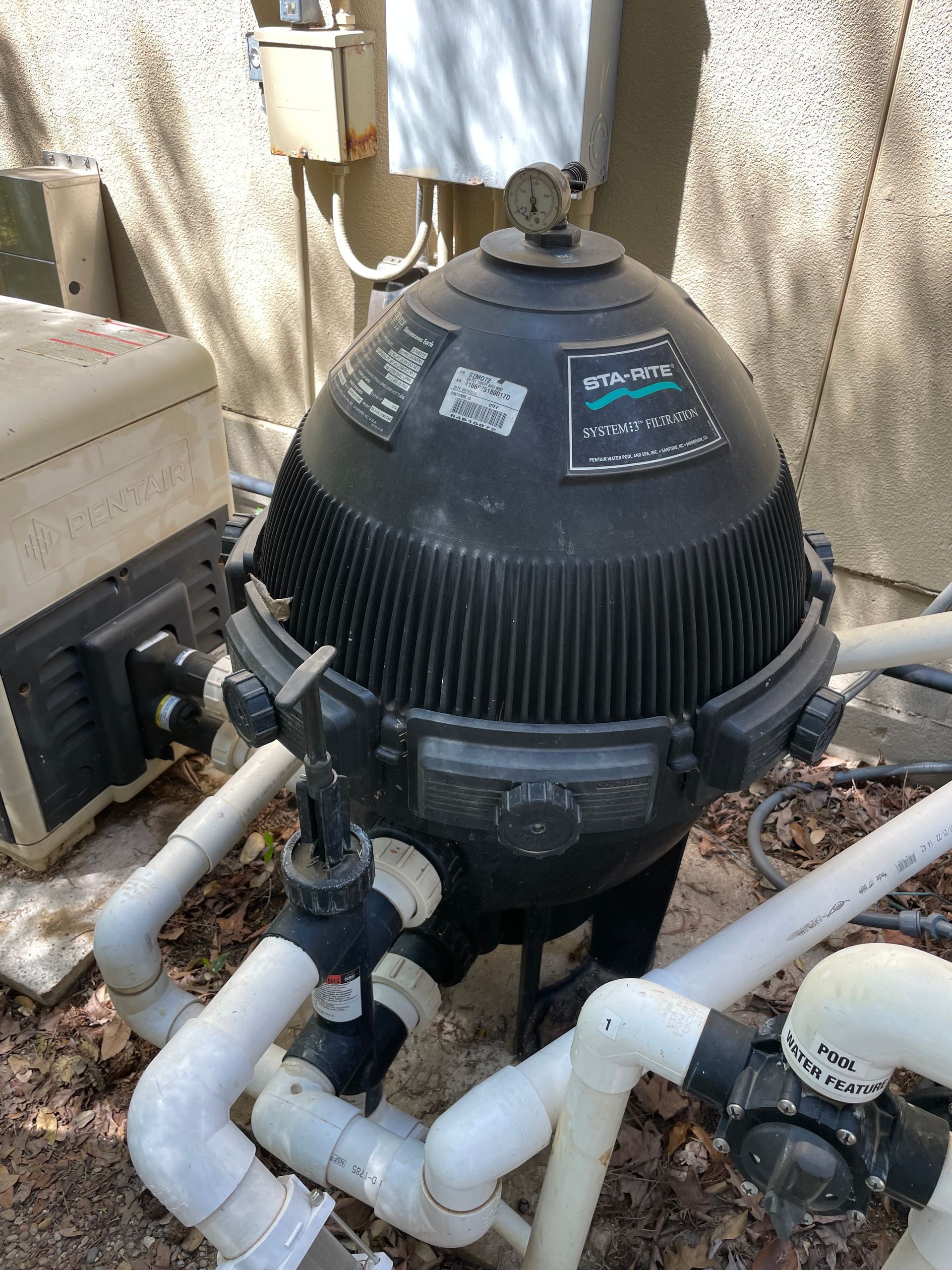 Black pool pump/filter unit with white pipes and valves outdoors against a beige wall
