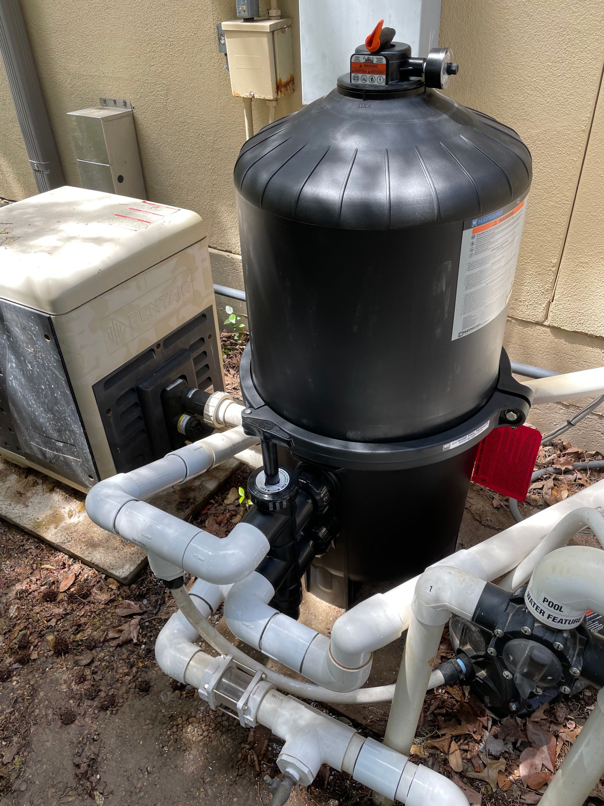 Black pool filter tank with white pipes and pump equipment beside a stucco wall outdoors