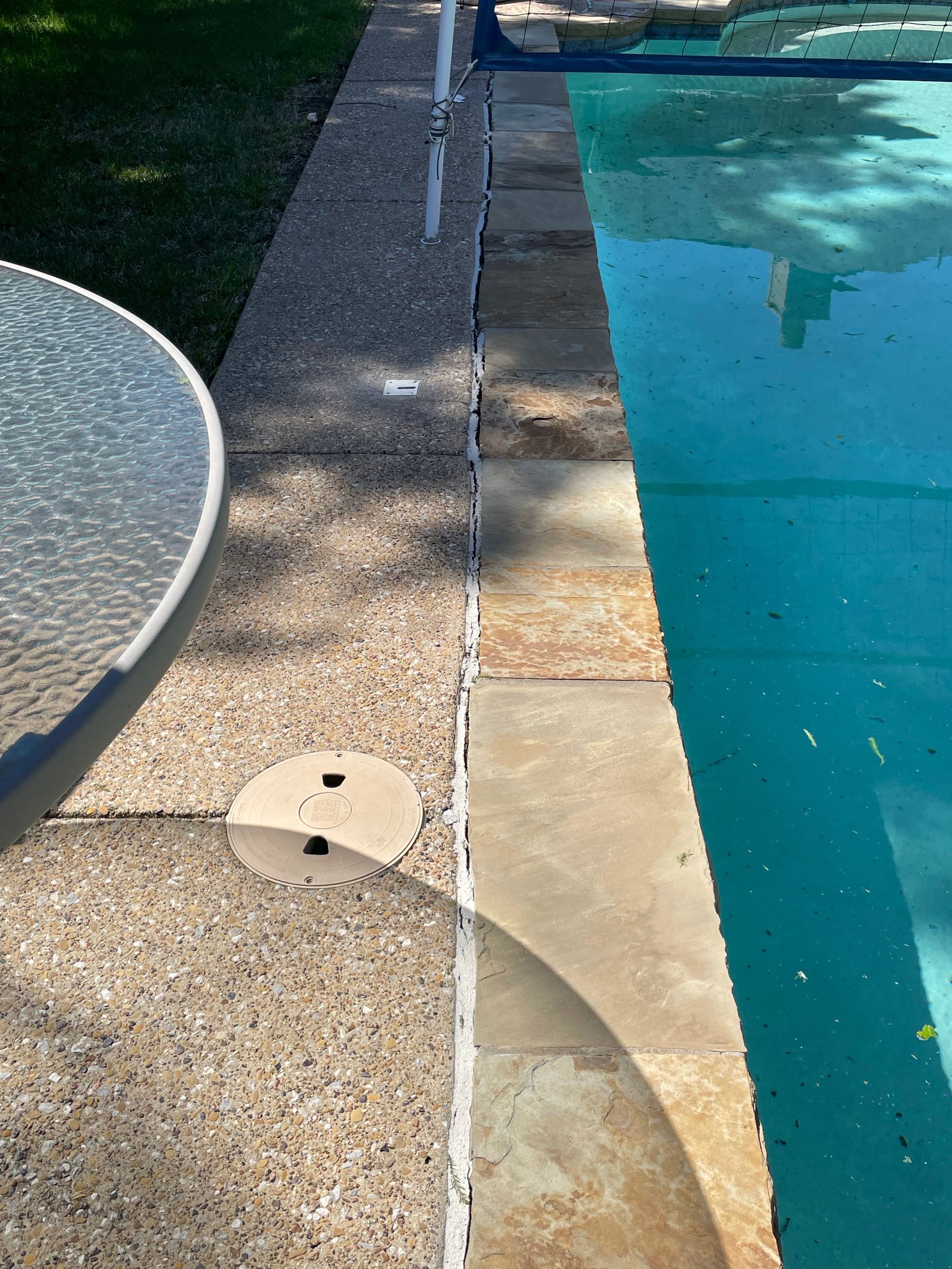 Swimming pool edge with wet concrete deck, drain line, and a round glass table beside the water