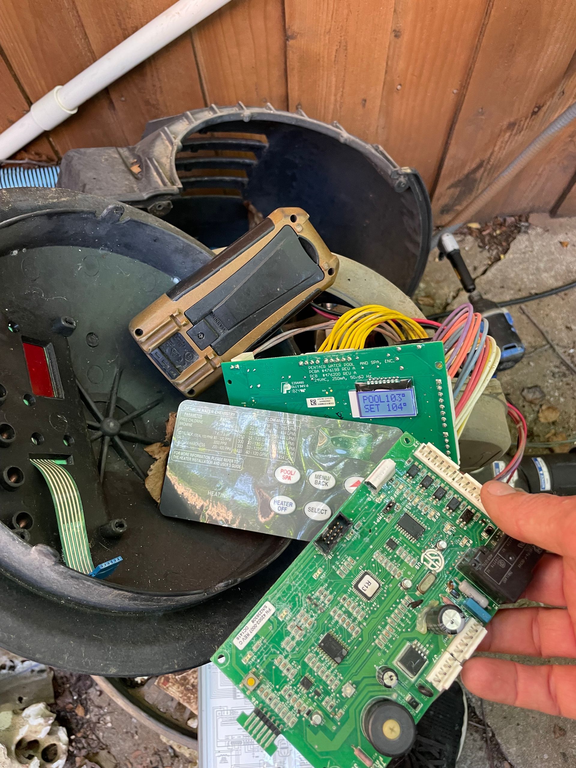 Hand holding a green circuit board over a pile of dismantled electronics and wires in a metal bin
