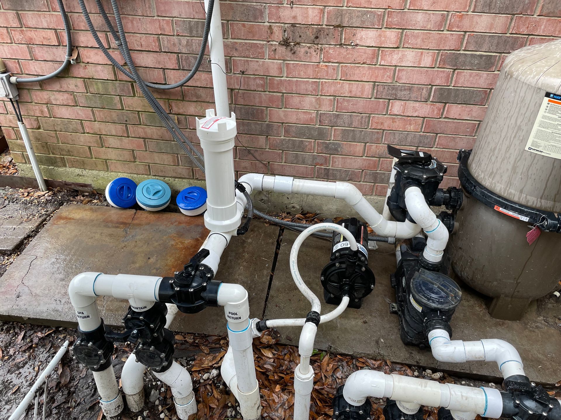 Pool plumbing with white PVC pipes and black pumps beside a brick wall.