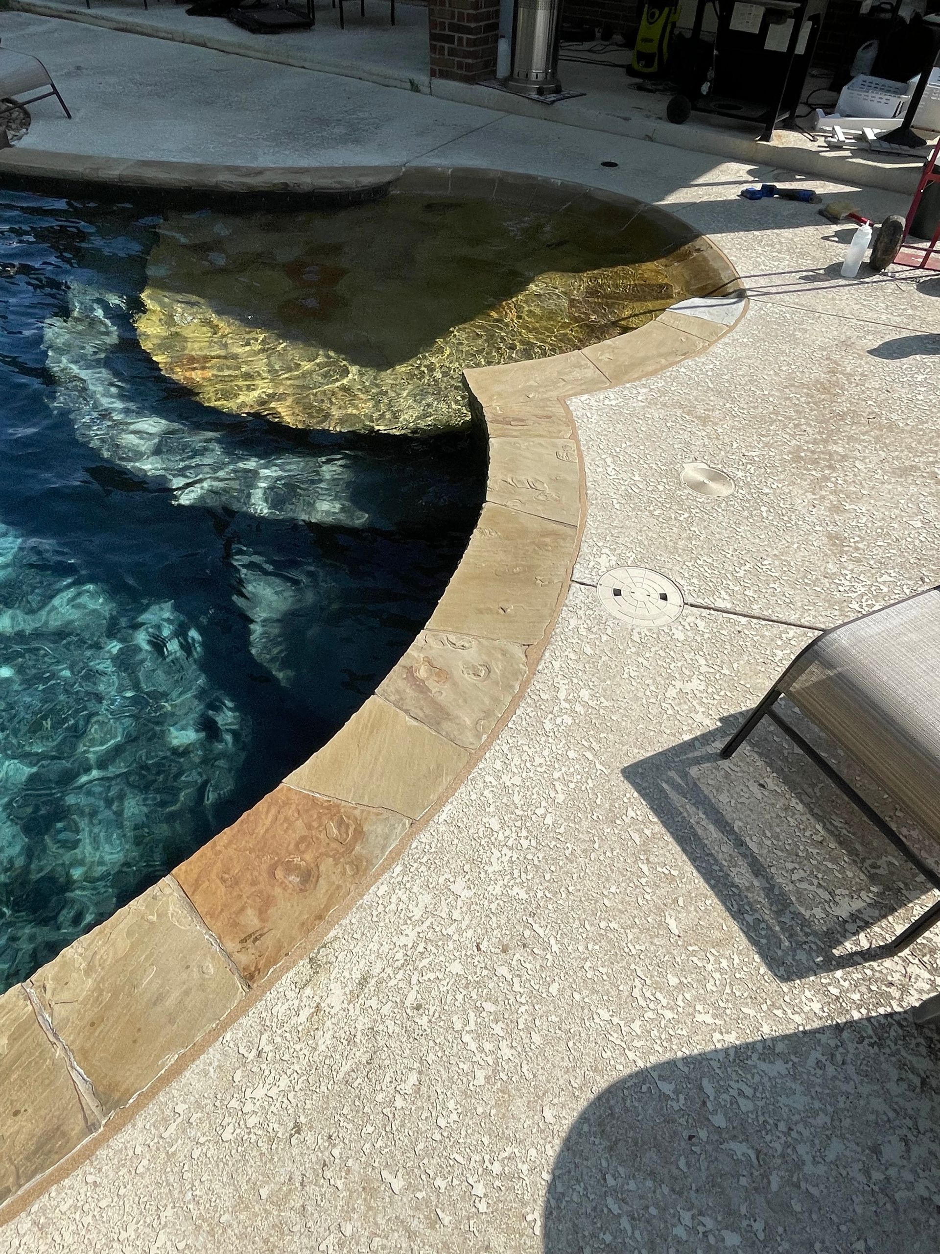 Curved swimming pool edge with tan stone coping, blue water, and a speckled patio beside it