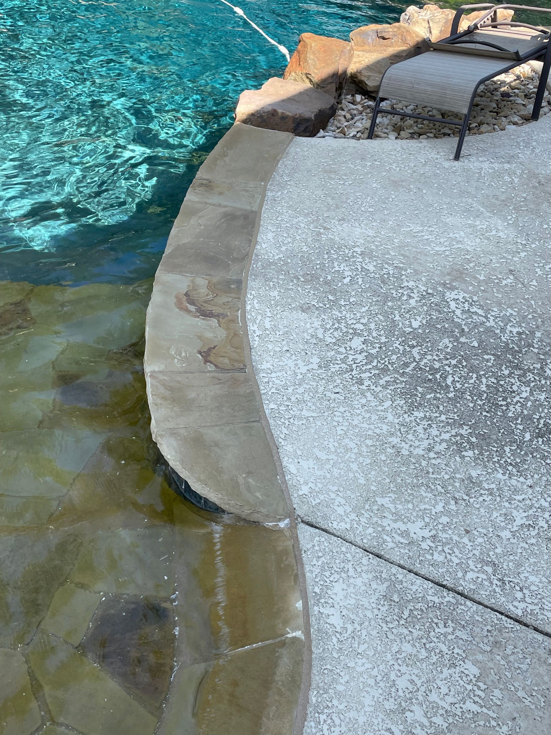 Curved concrete pool edge beside clear turquoise water and a rocky shoreline.
