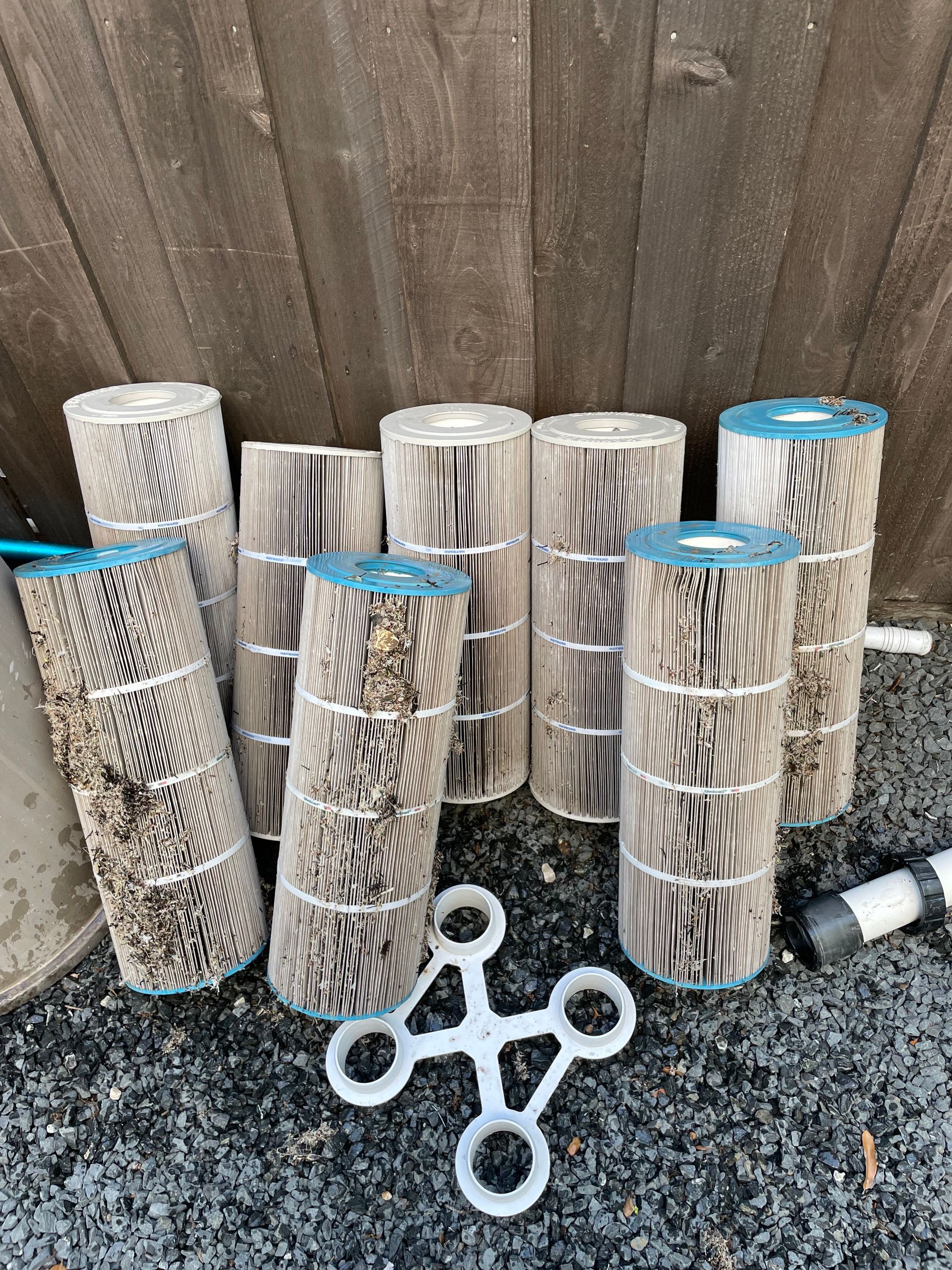 Rolled woven fence screens and white plastic garden stakes leaning against a wooden fence on gravel.