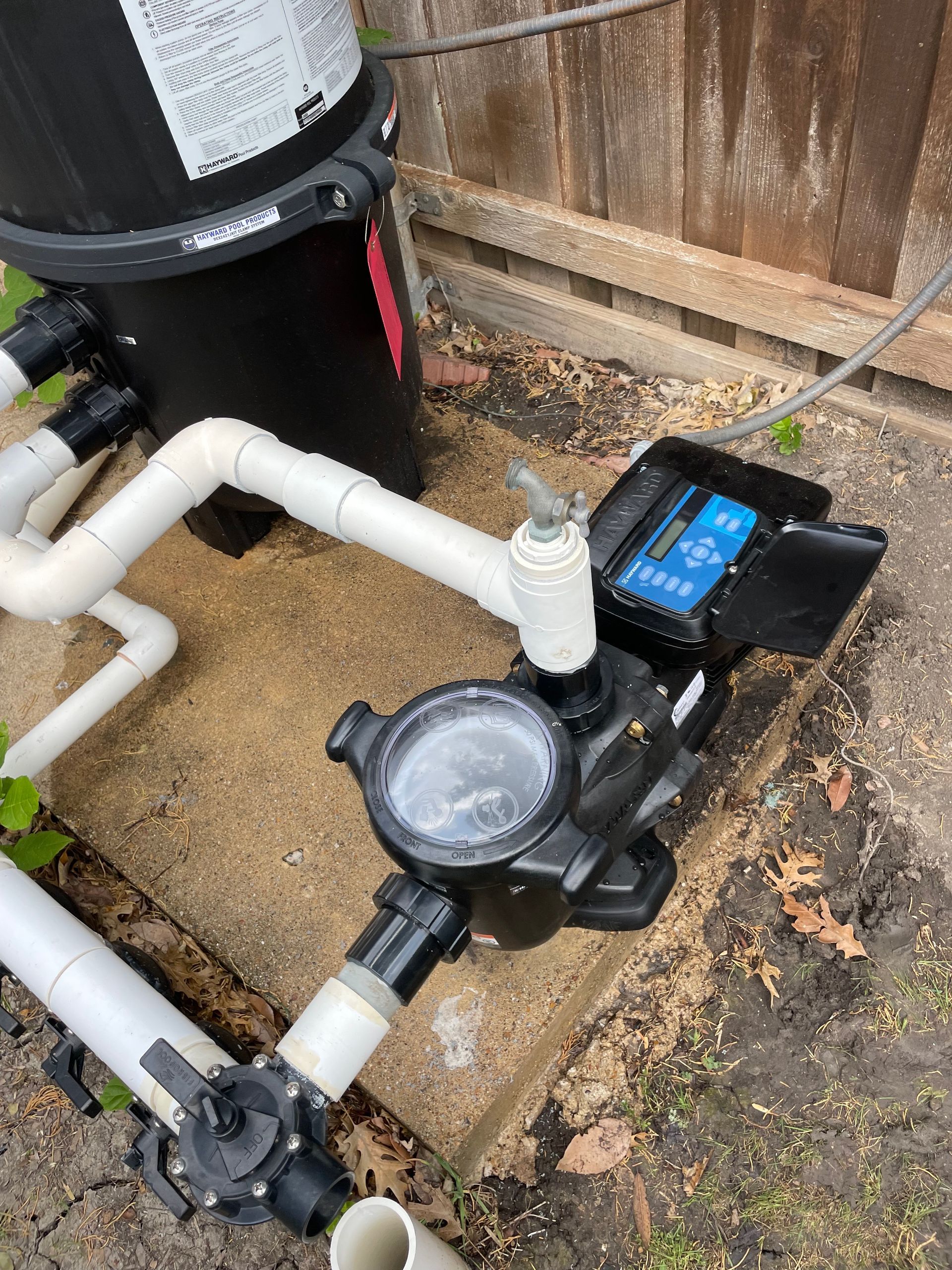 Pool pump and filter plumbing with black and white pipes beside a wooden fence.
