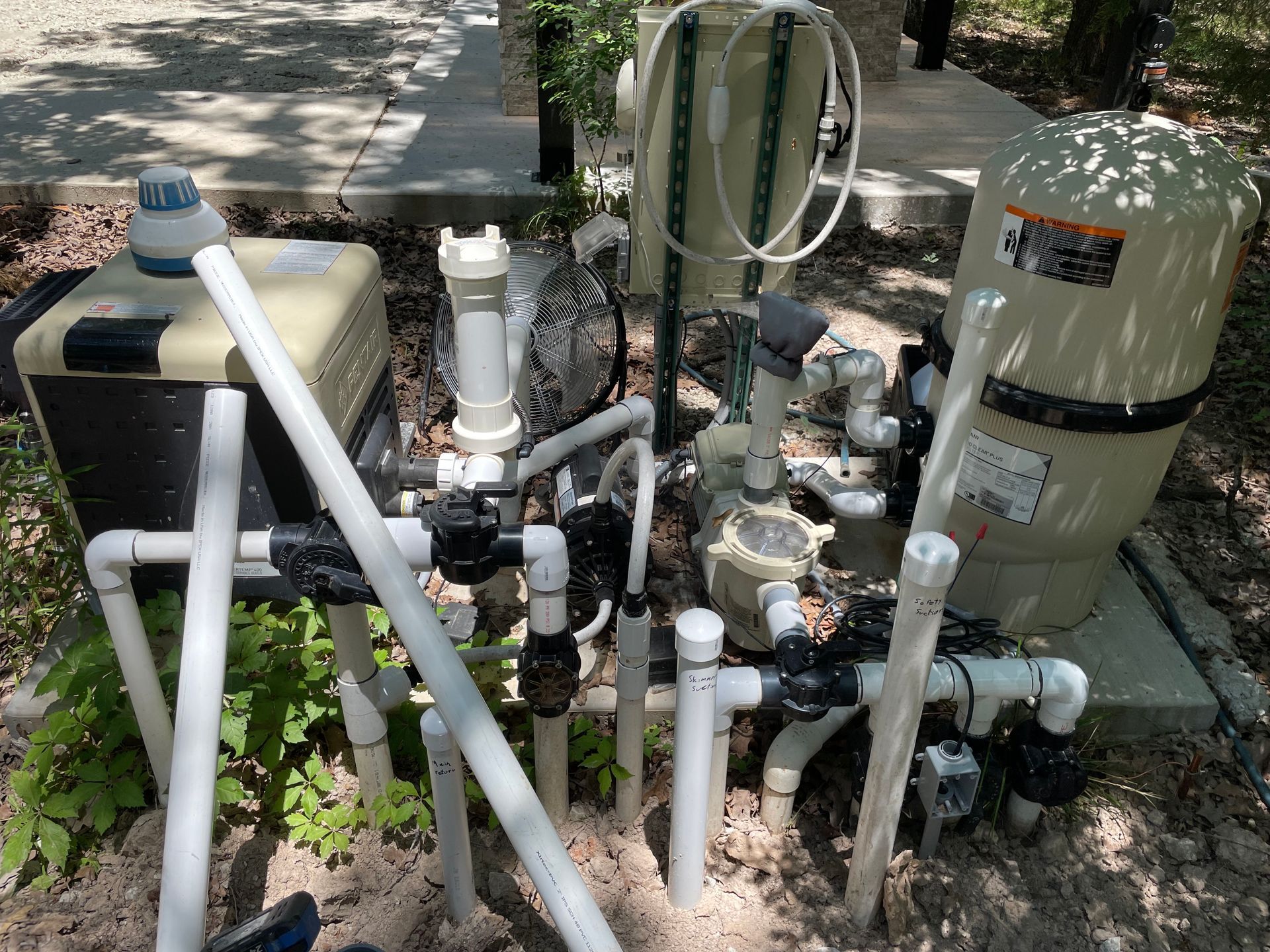 Outdoor pool equipment with pipes, pumps, and filters beside a concrete pad in a yard